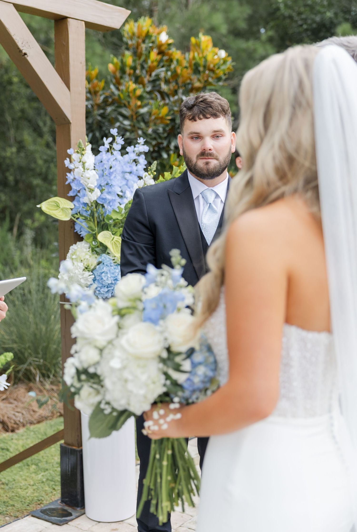 Groom in dark suit watching bride approach at outdoor wedding ceremony with blue floral arrangements.
