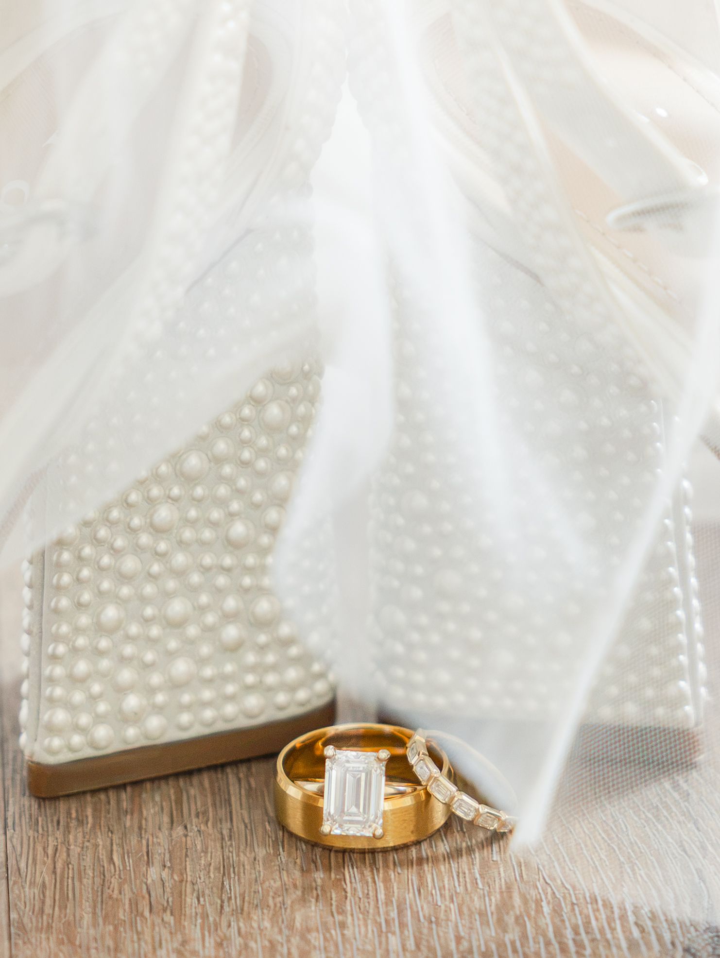 Gold wedding bands and emerald-cut diamond engagement ring displayed with pearl-studded heels and white bridal veil.