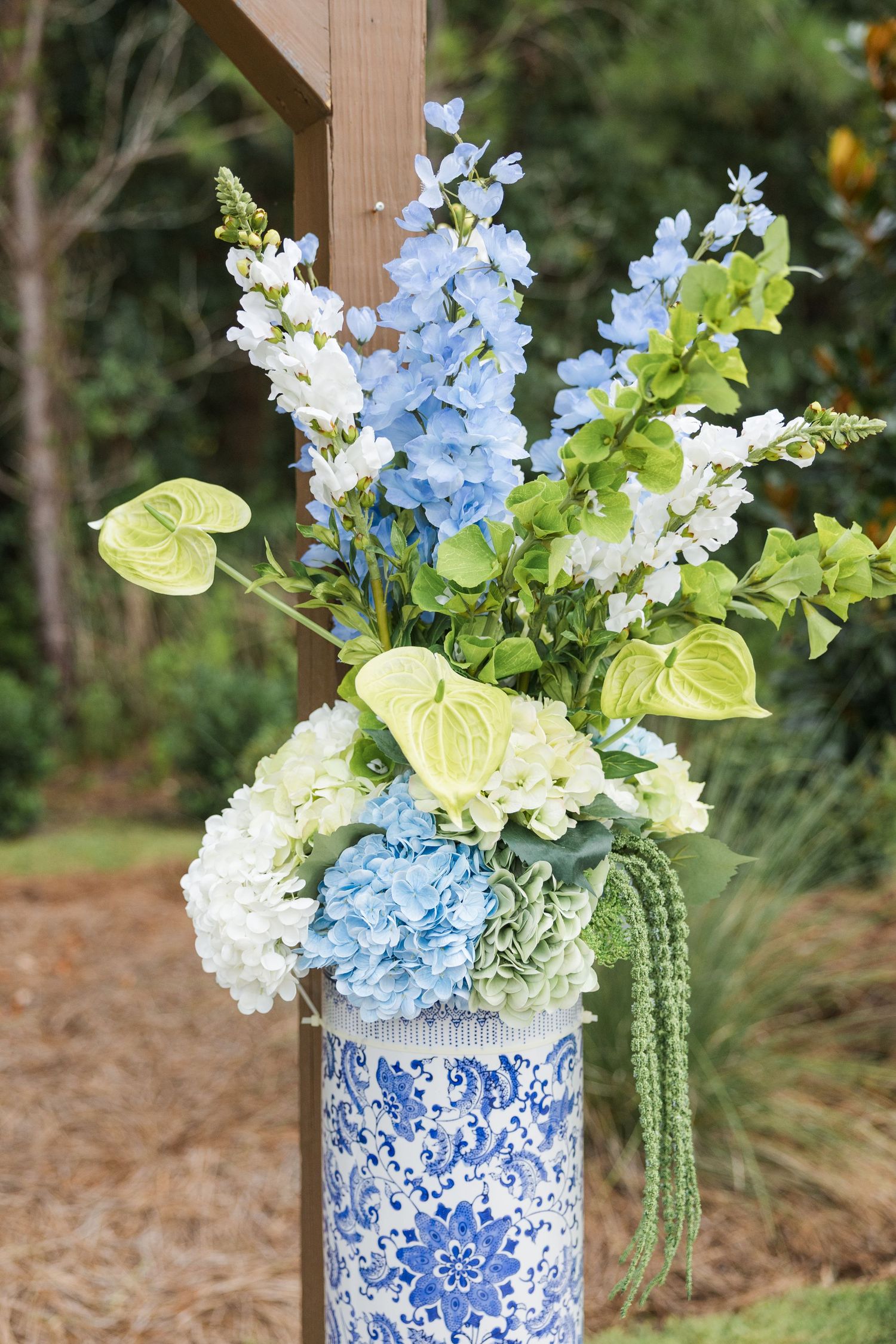 Blue and white chinoiserie vase with hydrangeas, delphiniums, and green anthurium at outdoor wedding ceremony.