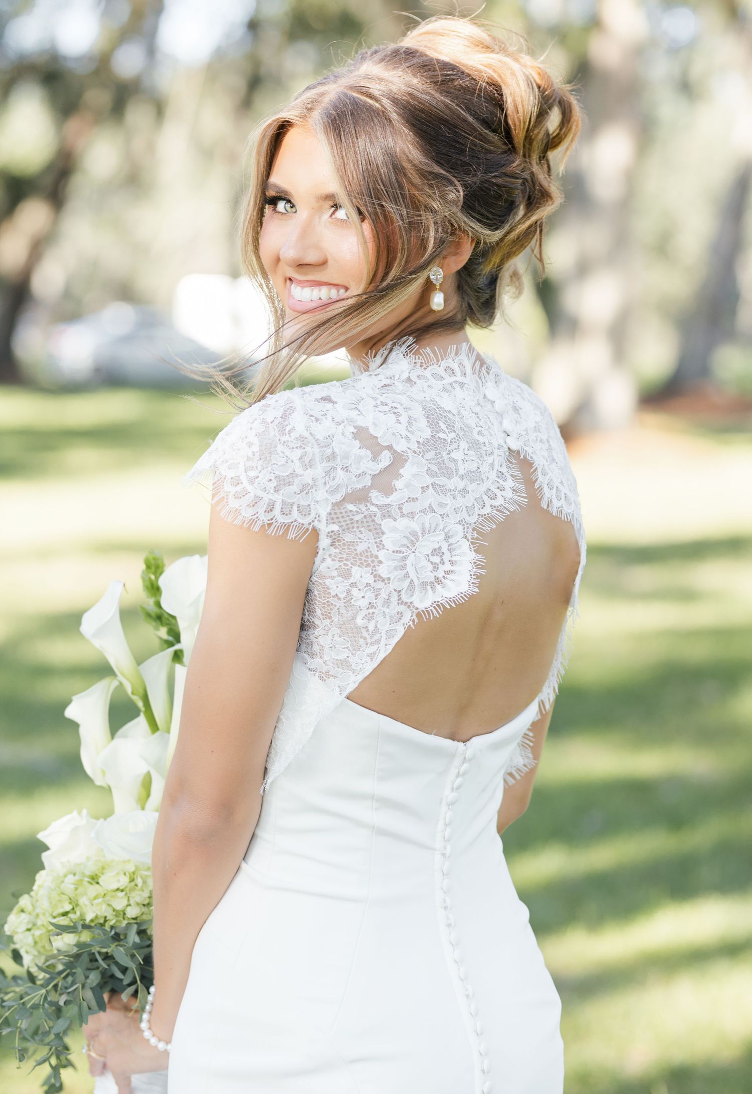 Smiling bride in a white lace back open keyhole wedding gown holding a bouquet of white flowers outdoors.