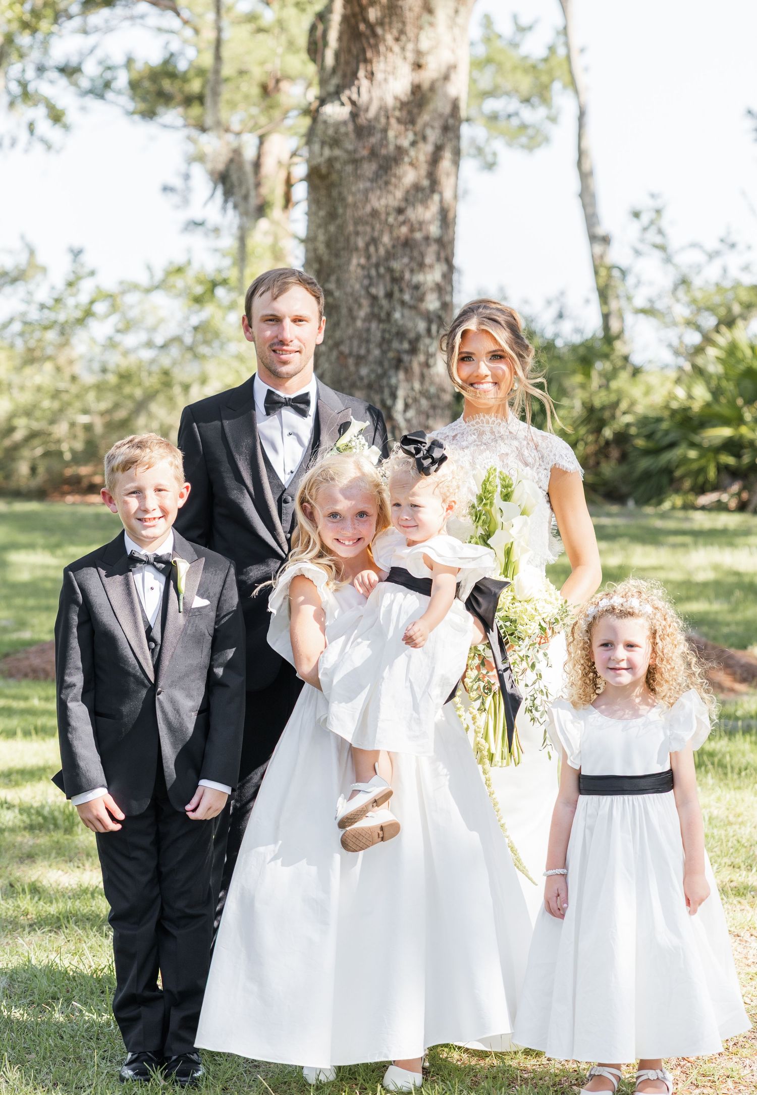 Bride and groom pose with four children in formal attire outdoors near a large tree on their wedding day.