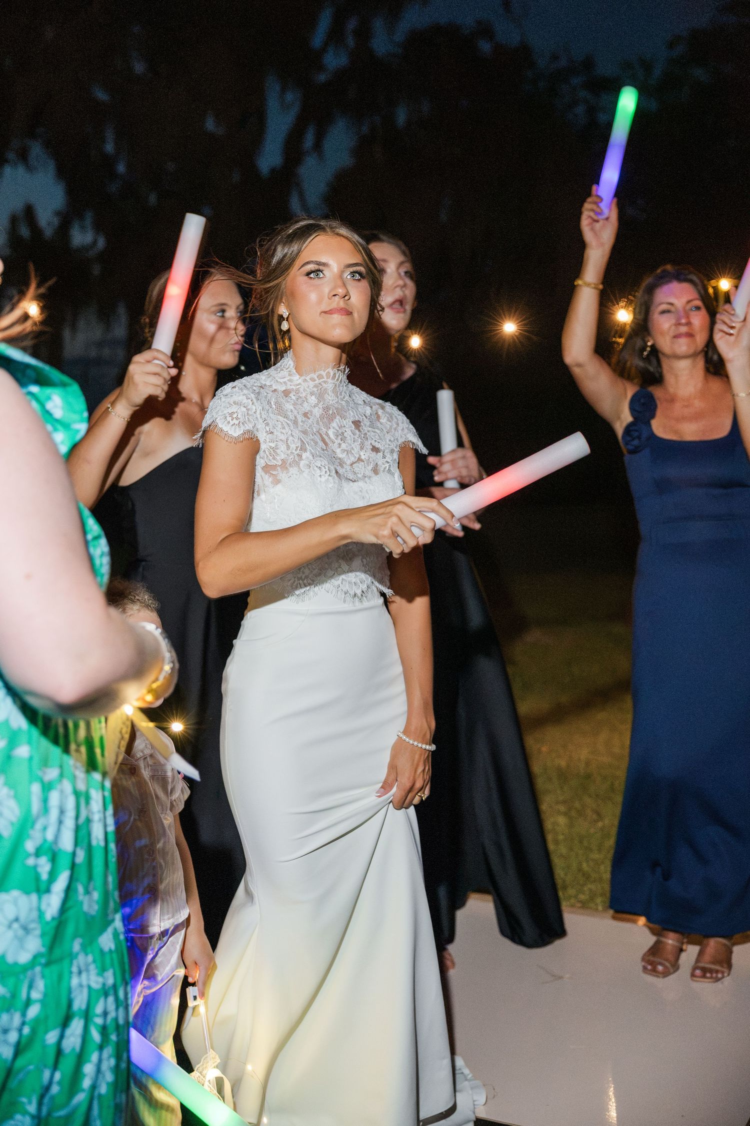 Bride in white lace gown holds glowing foam stick at outdoor wedding reception at night.