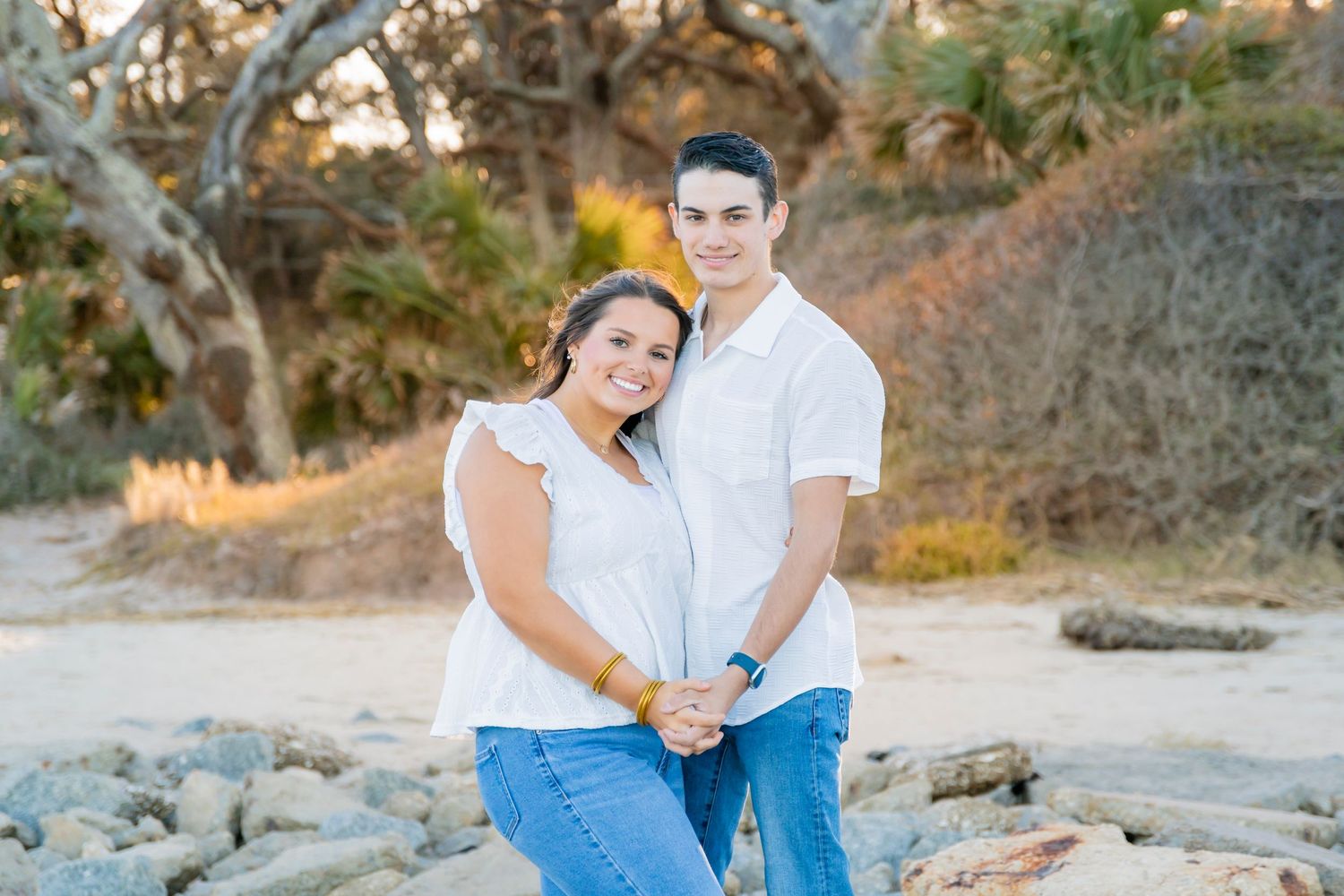 Casual beach portrait session in white and blue outfits against rocky coastline.