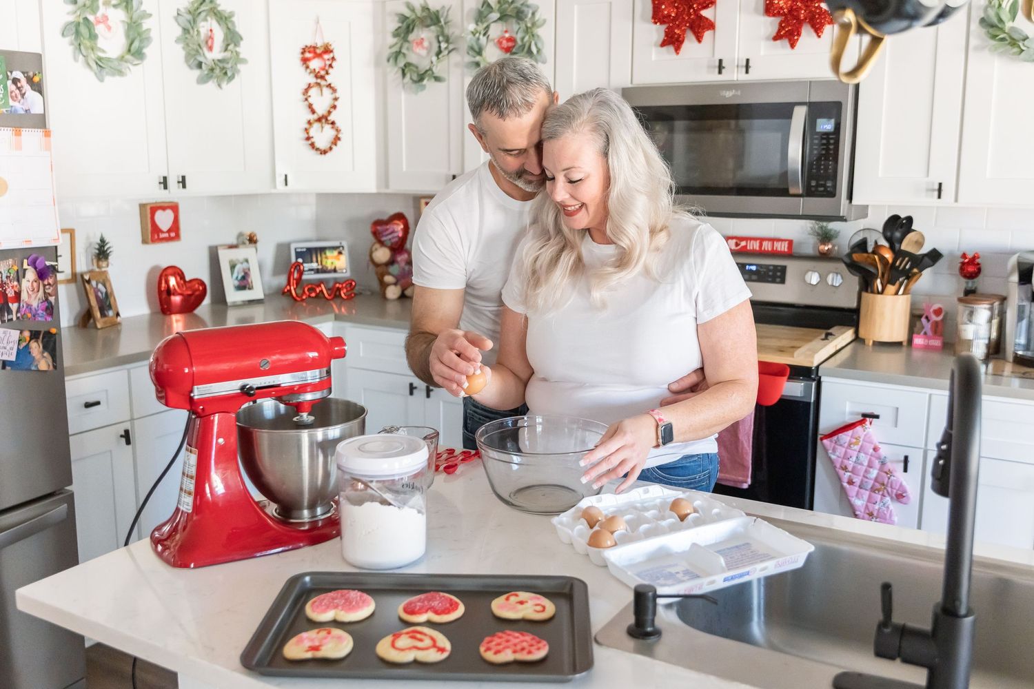 A couple makes Christmas cookies together in a kitchen with a red stand mixer and holiday decorations.
