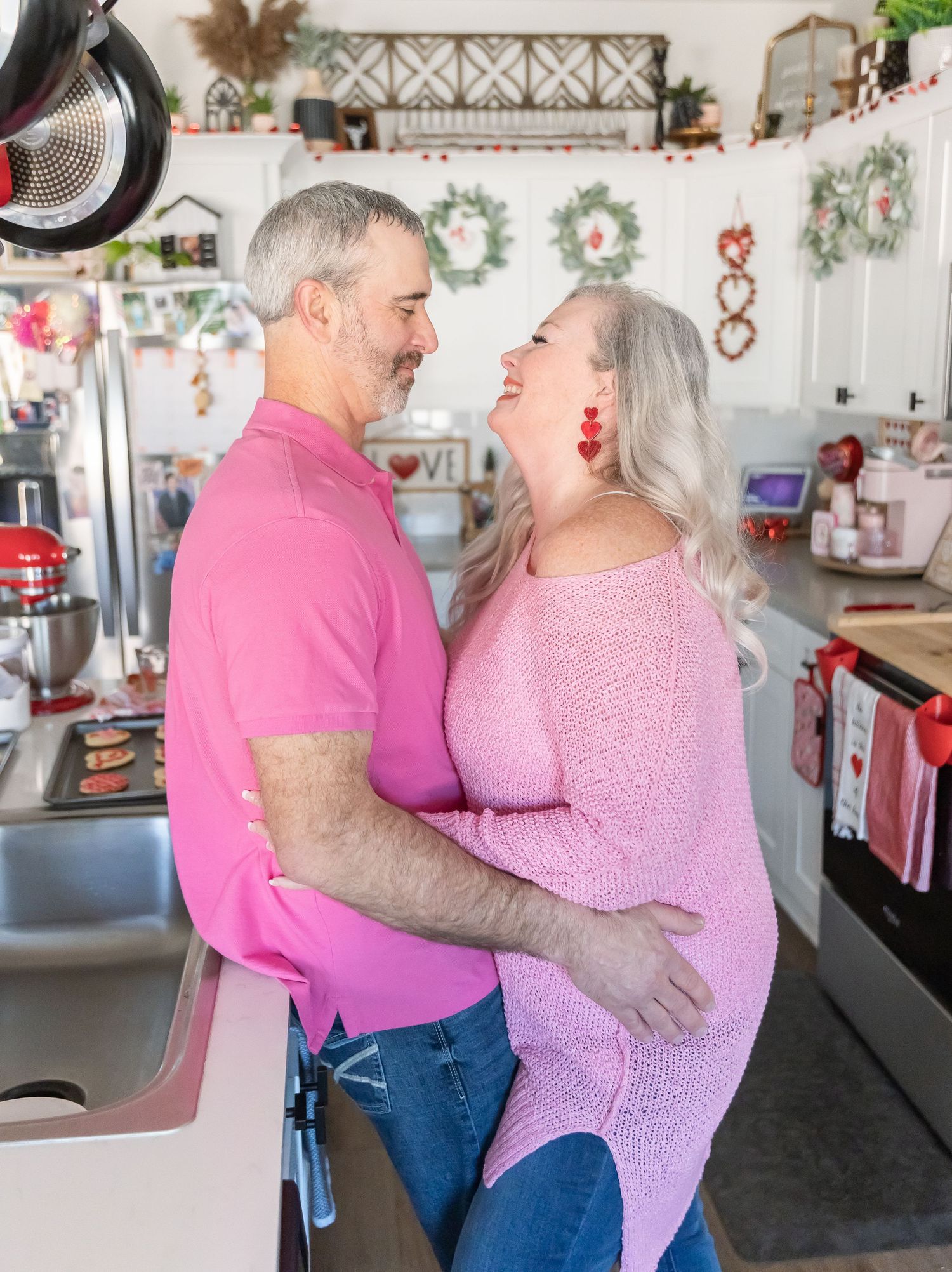 A couple dressed in matching pink outfits laughs together in their kitchen with holiday decor.