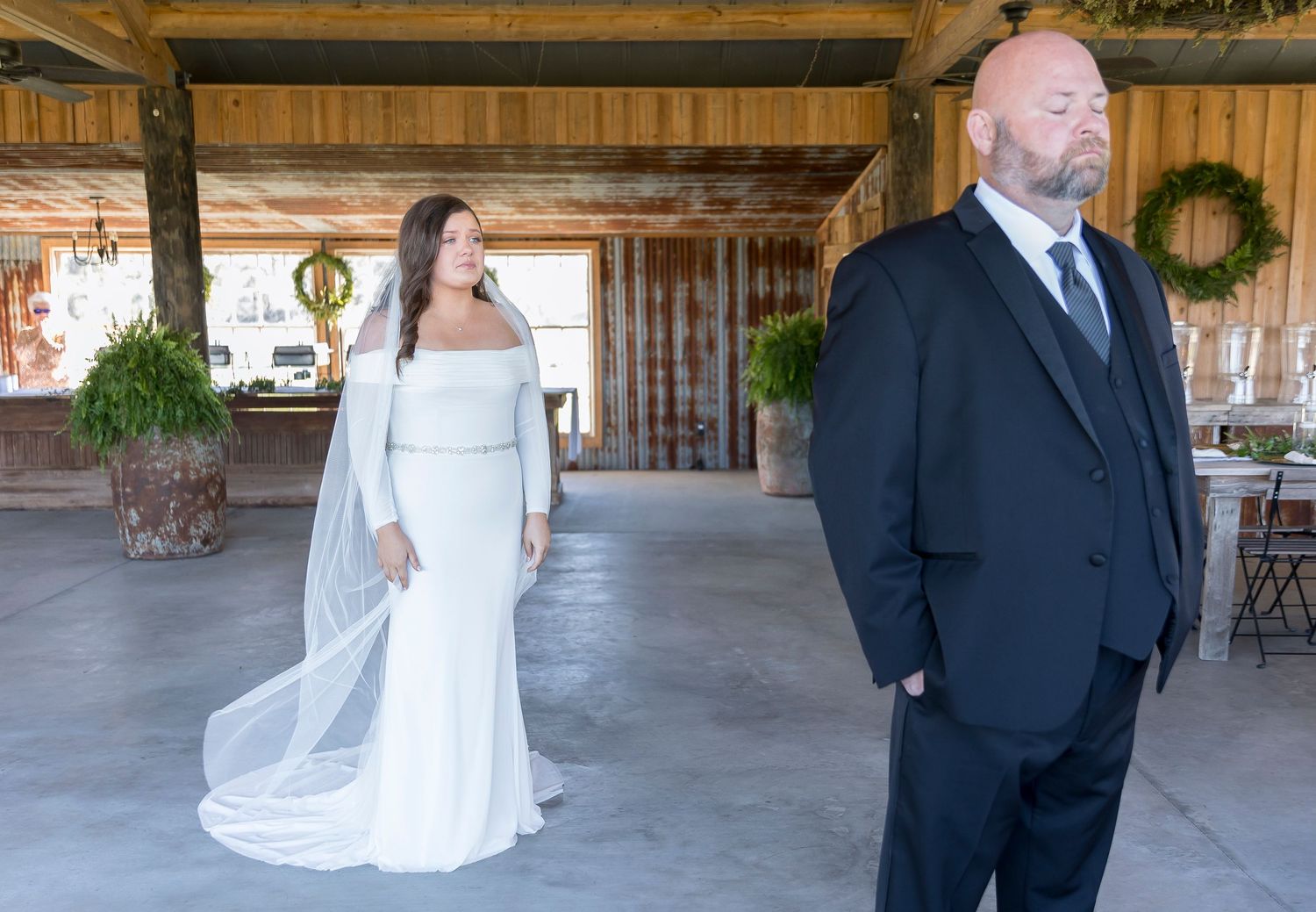 A couple in wedding attire stands apart in a rustic covered venue with wooden beams.