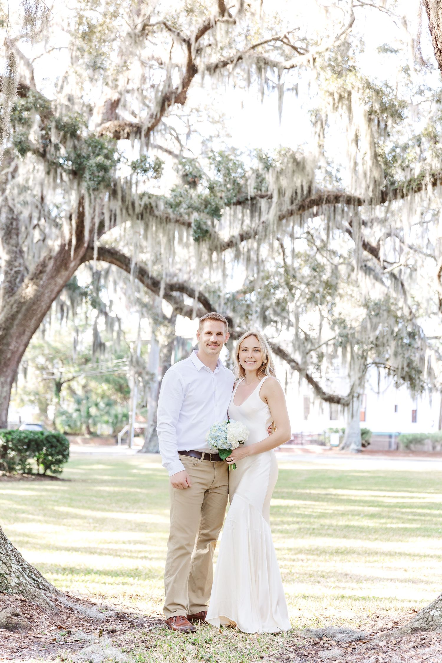 Newlywed couple poses under a majestic Spanish moss-covered oak tree in a sunny park setting.