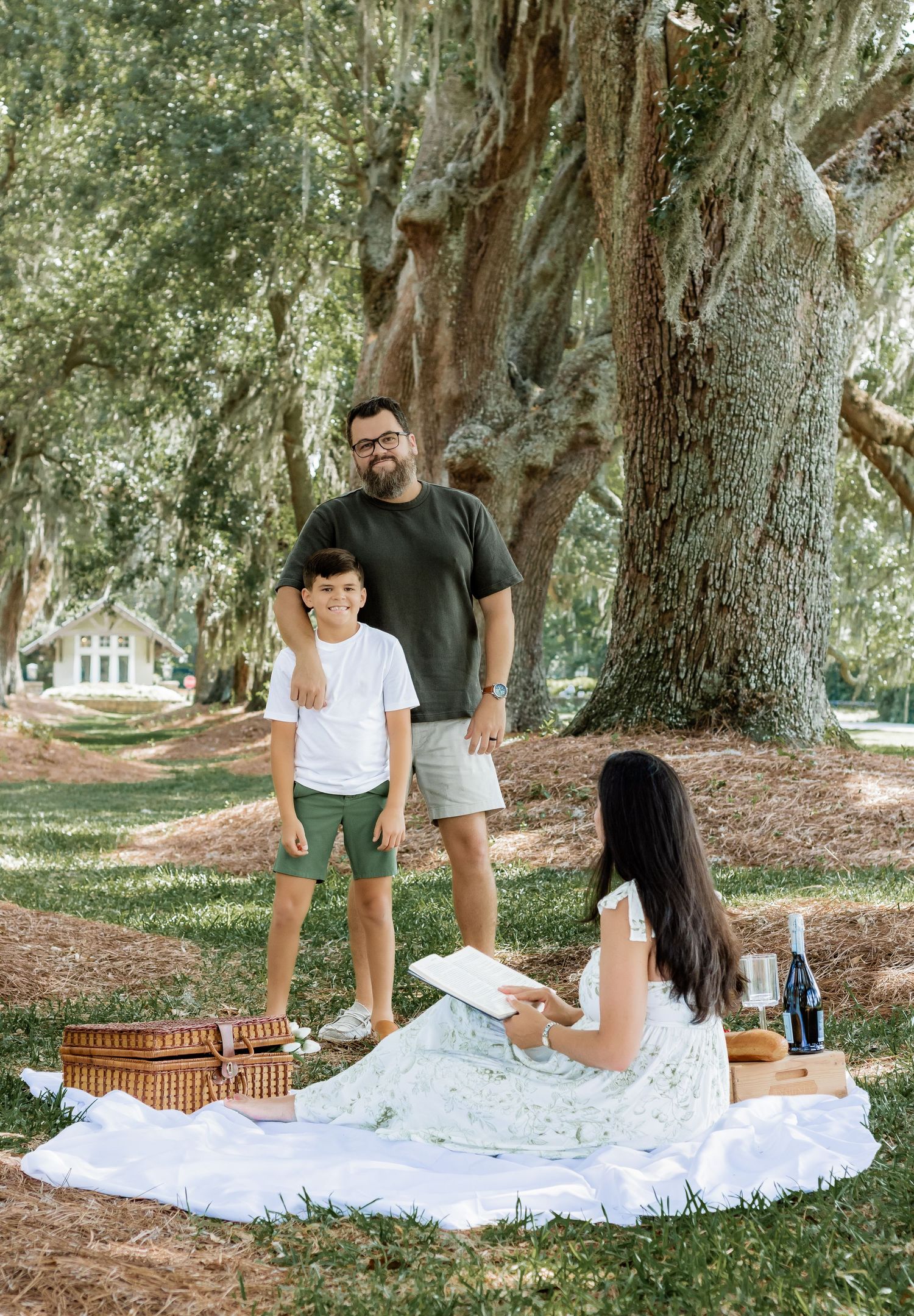 A relaxed family gathering at a picnic with a wicker basket under shady oak trees.