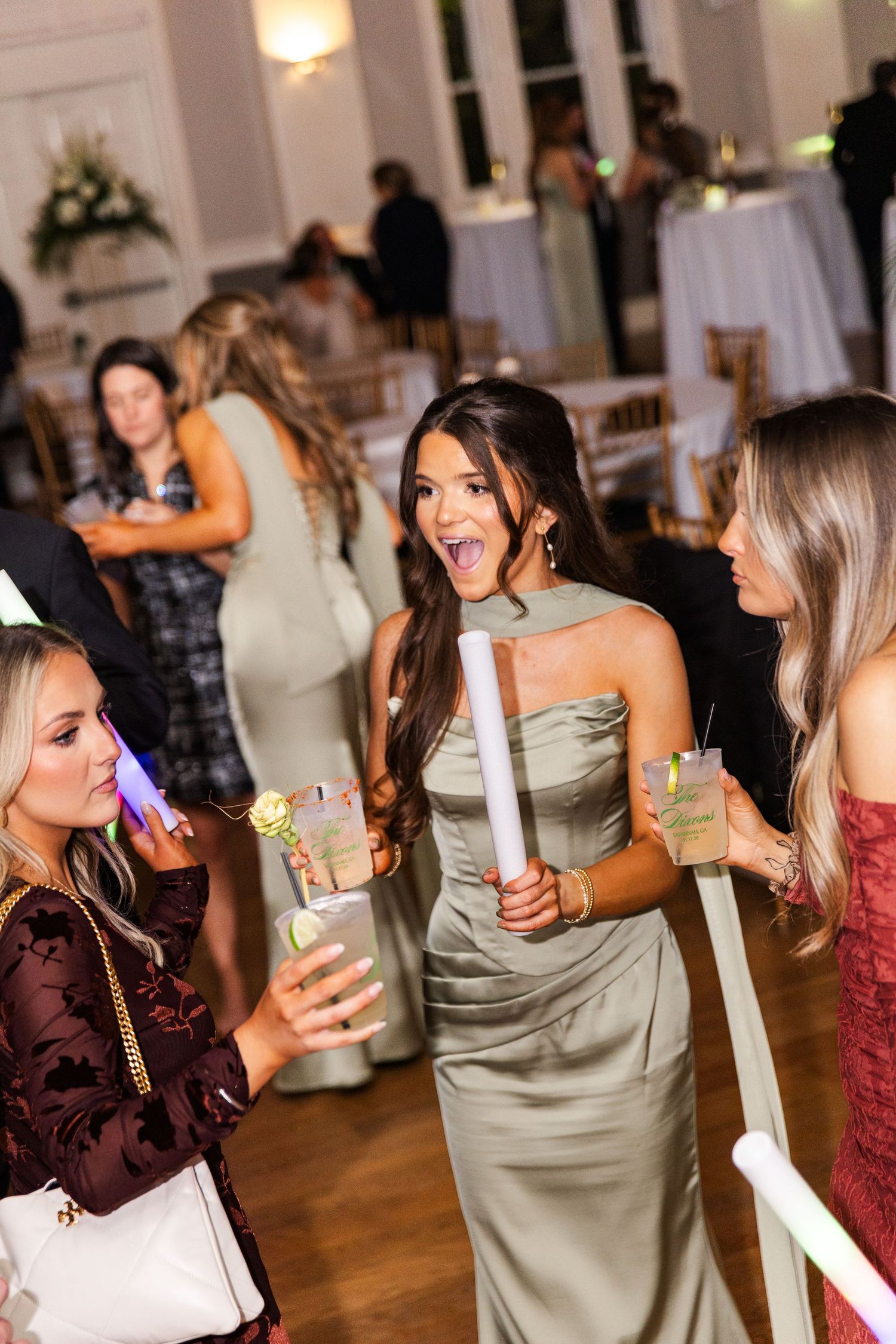 Bridesmaid in sage green satin dress holding glow stick and drink at wedding reception.