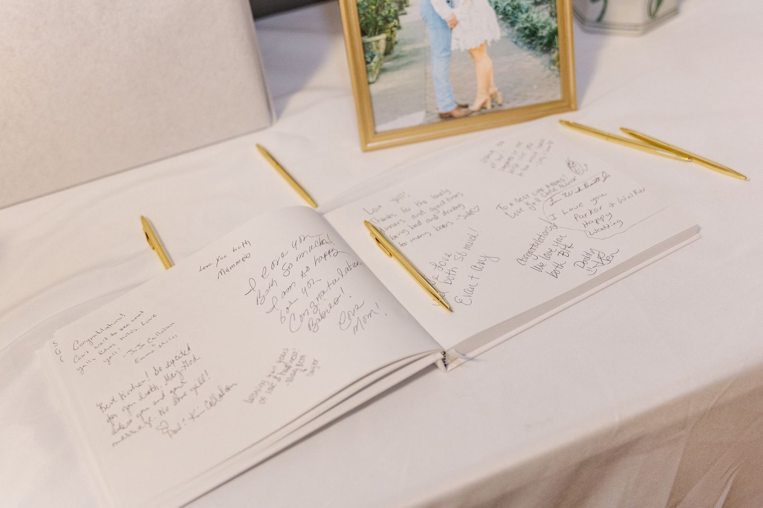 Wedding guest book open on a white table with gold pens and a framed couple photo in the background.