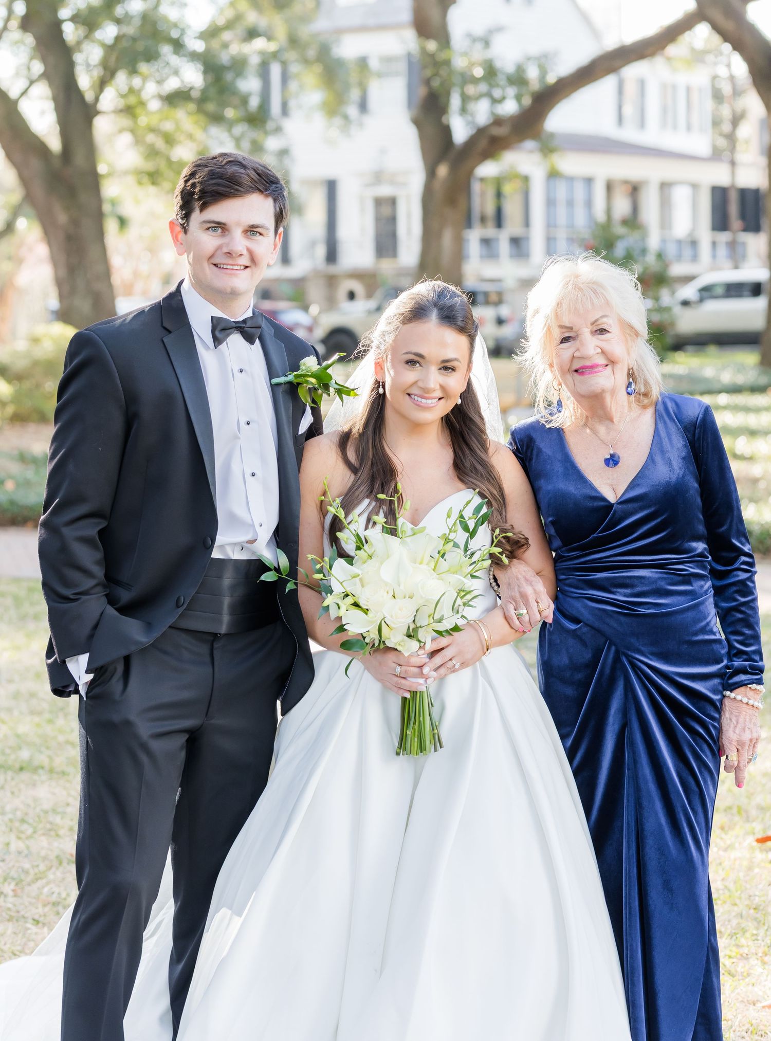 Bride in white gown holds bouquet flanked by groom in tuxedo and elderly woman in navy dress at outdoor wedding.