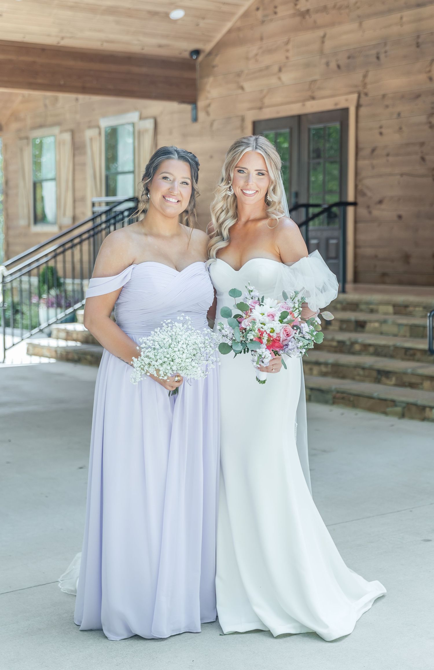 A bride in white and bridesmaid in lavender pose together on a wooden porch holding floral bouquets.
