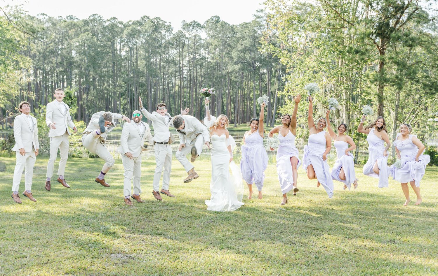 Wedding party jumping and celebrating on grass with forest backdrop.