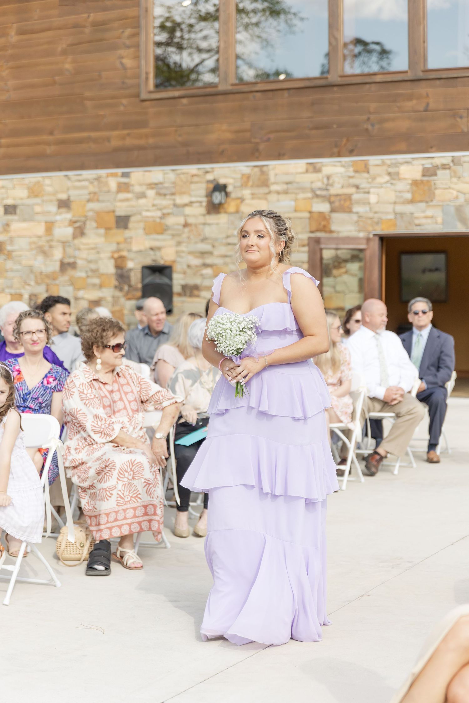 Bridesmaid in tiered lavender gown carries bouquet past seated wedding guests.