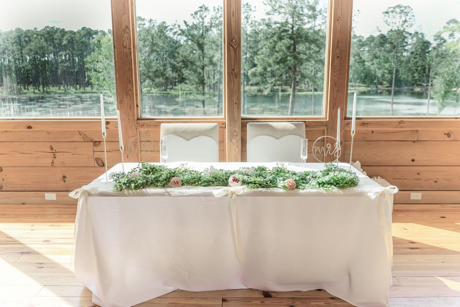 Rustic wedding reception table setup with white linens and greenery against large windows.