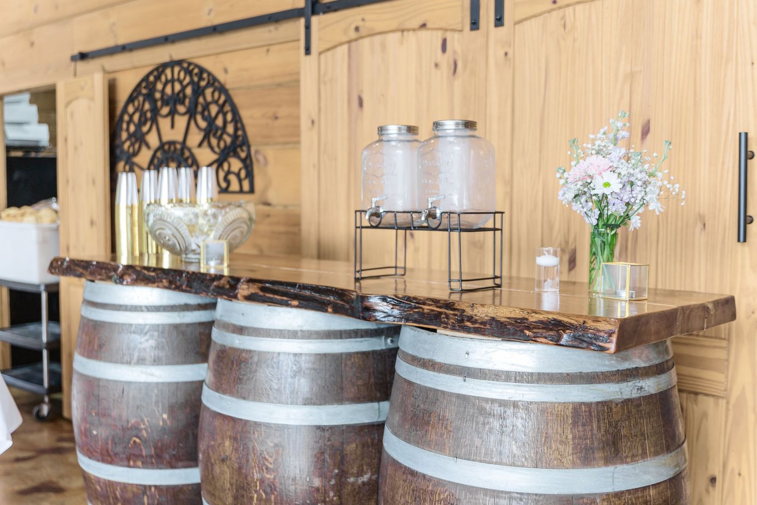 Rustic wedding bar setup with wine barrels and drink dispensers against wooden wall.