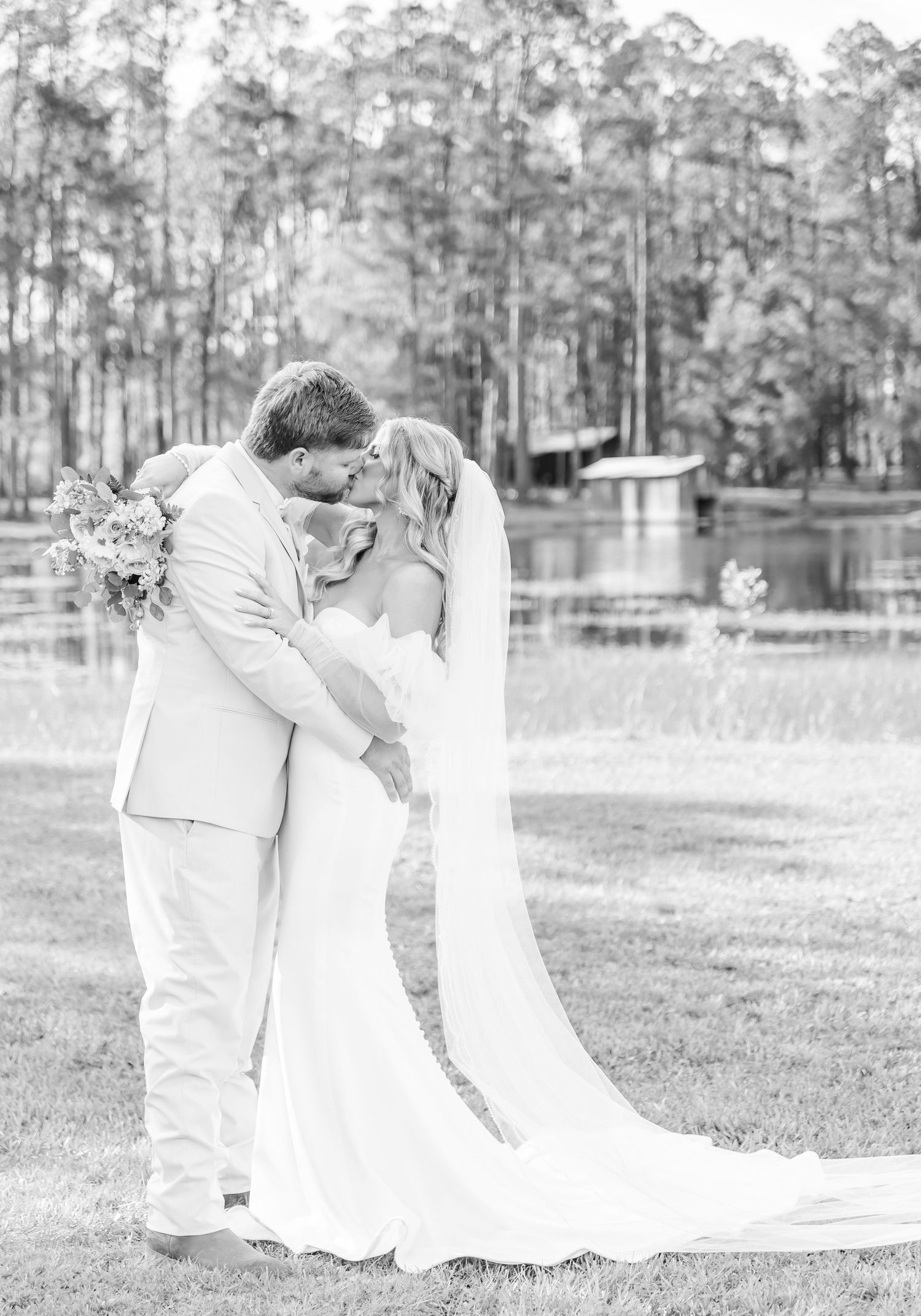 A romantic wedding couple shares a tender moment in front of a lake surrounded by tall pine trees.