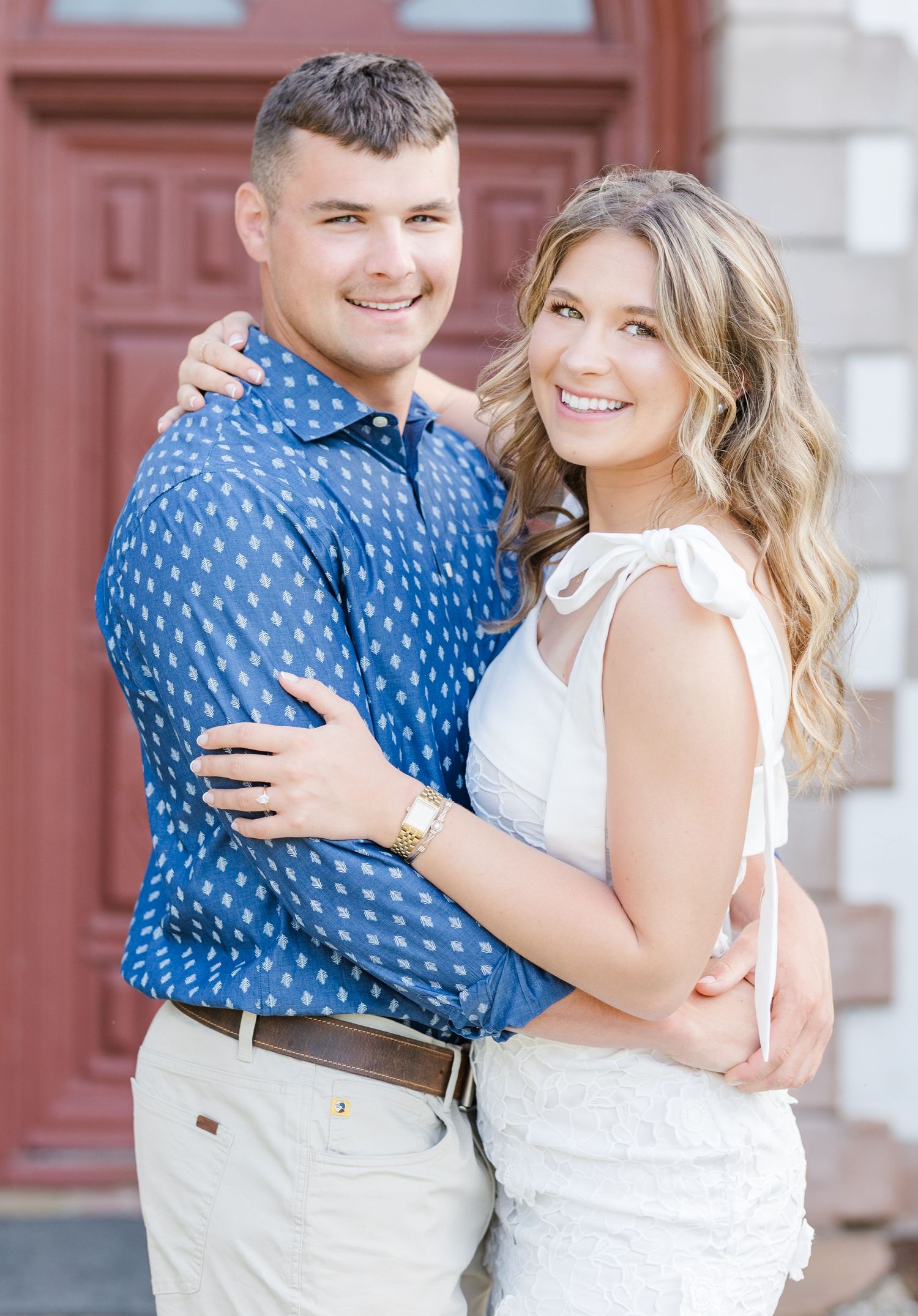 Smiling couple embracing outdoors, man in blue patterned shirt and woman in white dress, engagement photo.