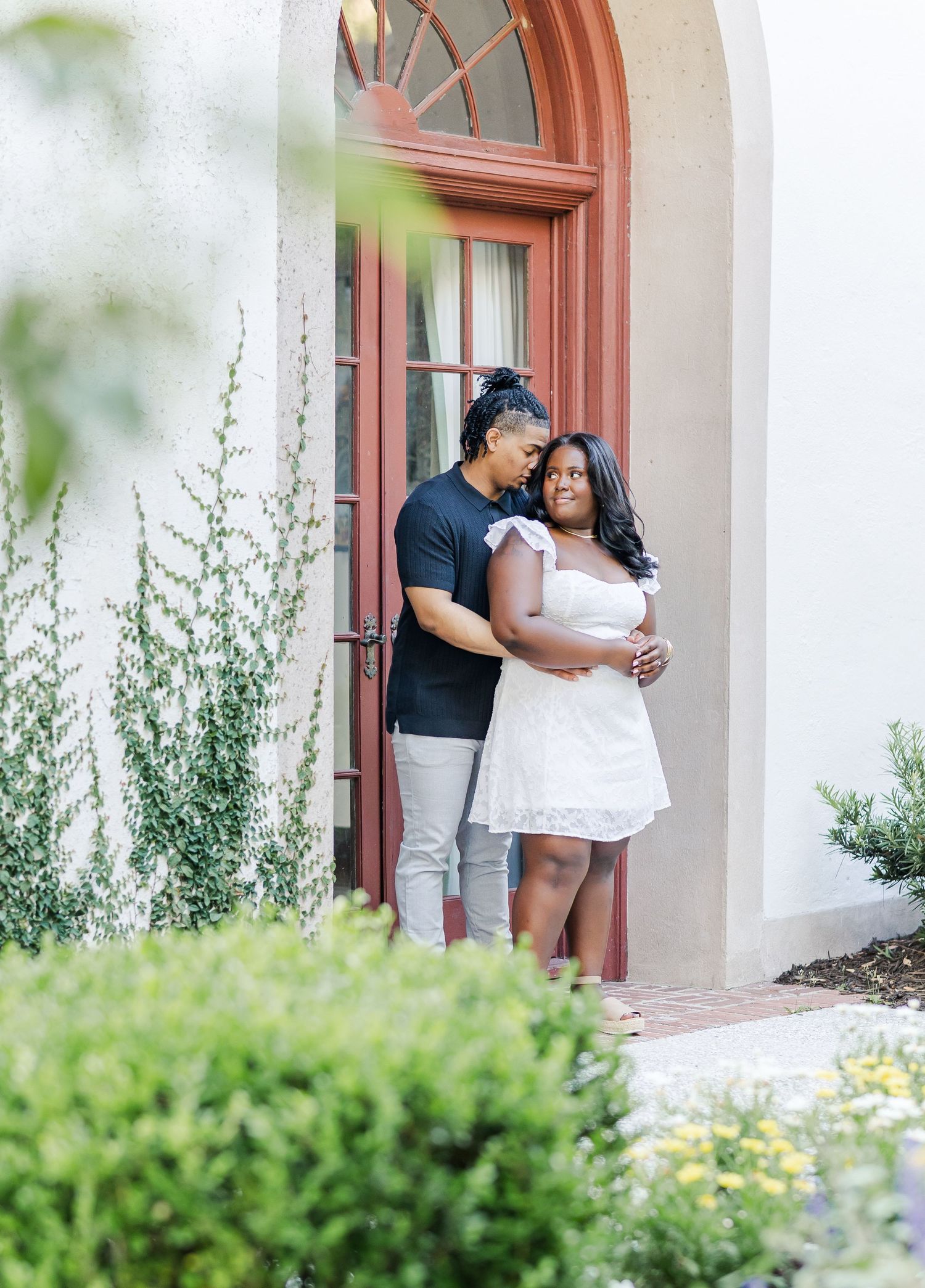 A couple embraces in a doorway, the man in black hugging the woman in a white dress from behind.