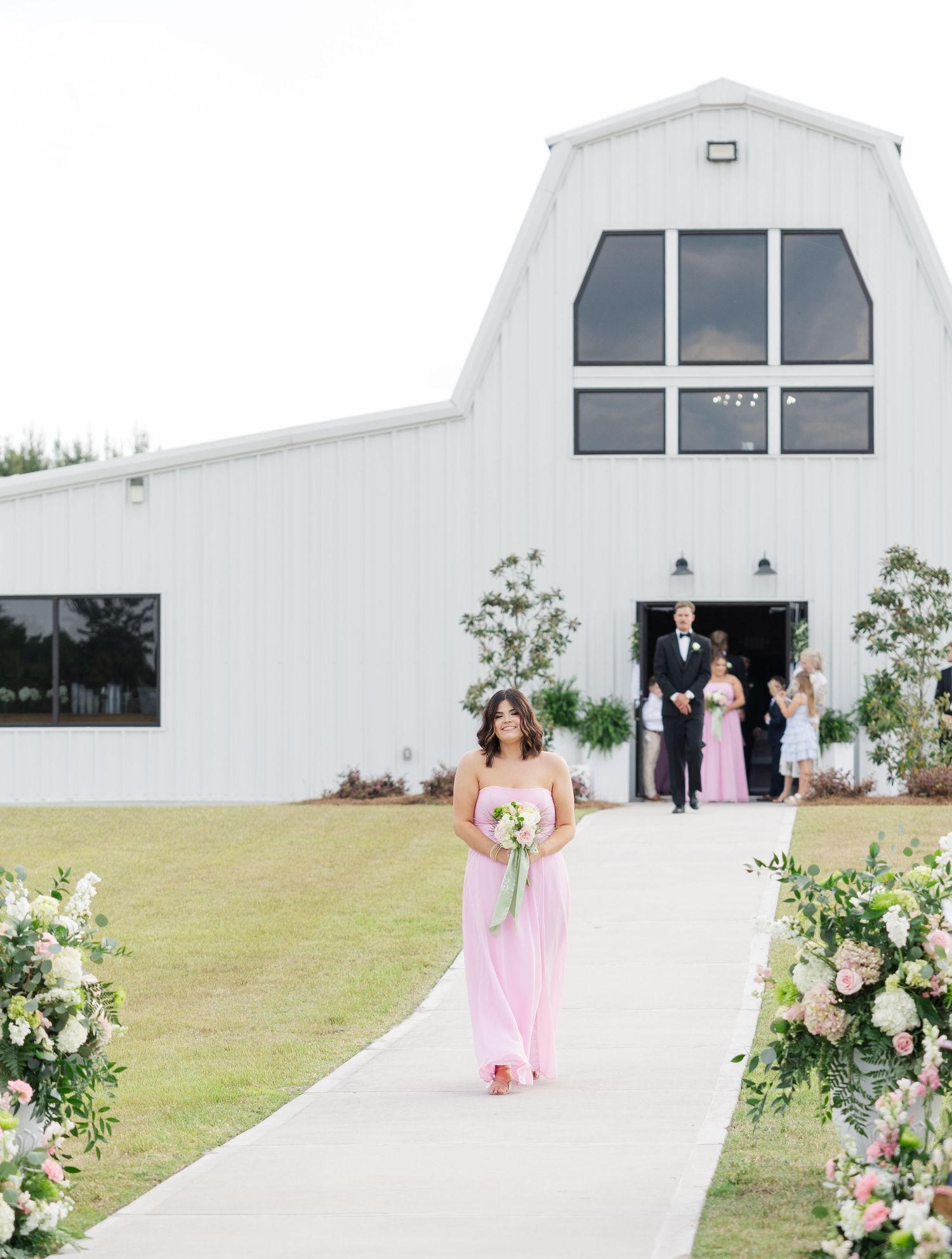 Bridesmaid in pink gown walks down outdoor aisle toward white barn wedding venue.