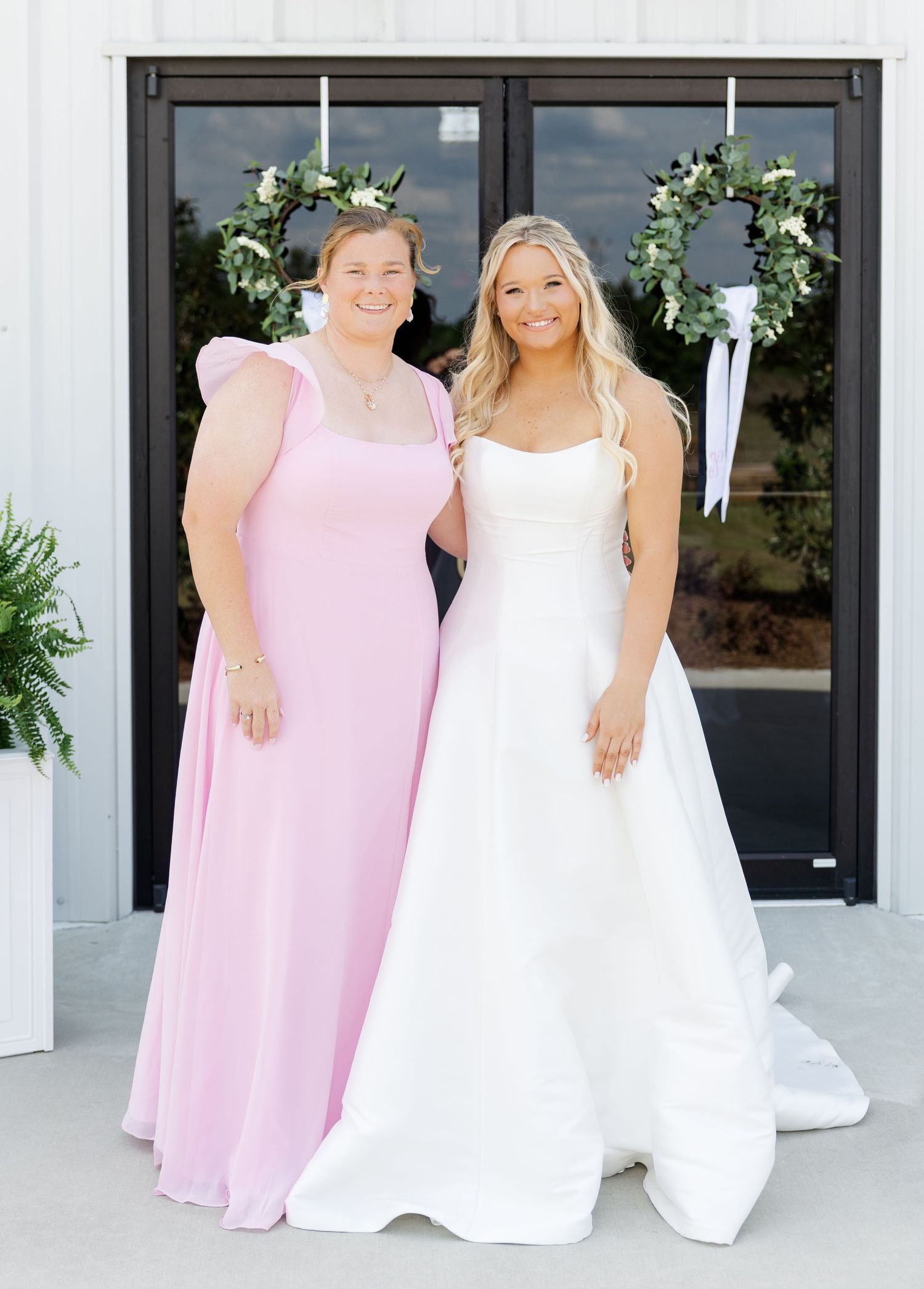 Bride in white gown poses with bridesmaid in pink dress before double doors adorned with greenery wreaths.