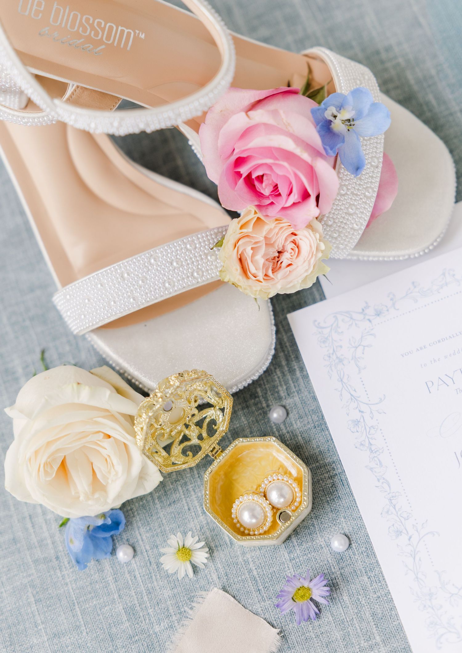 White bridal heels adorned with pearls and pink roses beside pearl earrings in a gold box and delicate blue flowers.