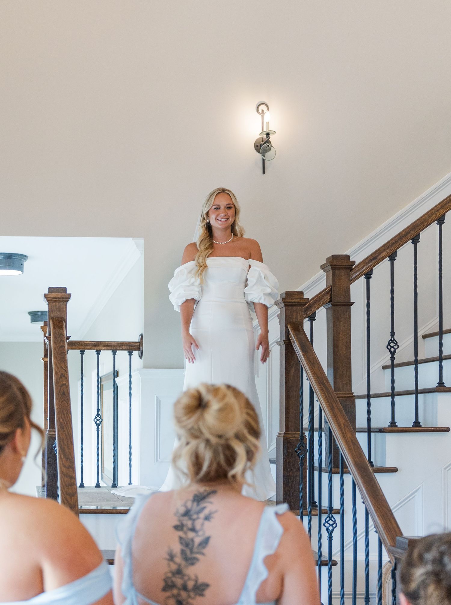Bride in white off-shoulder puff sleeve gown stands on staircase, smiling at bridesmaids below in elegant home.