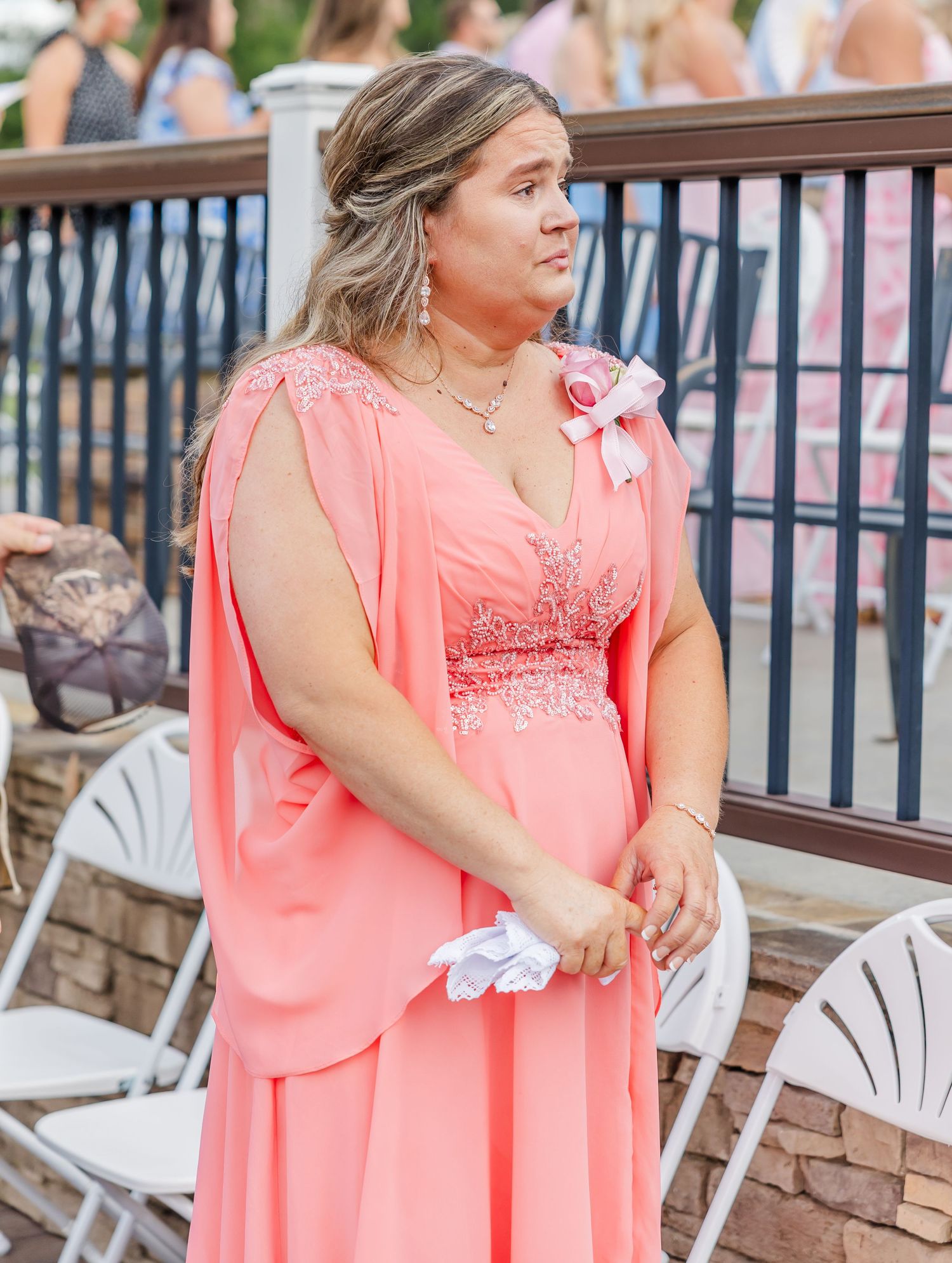 A bridesmaid in a coral pink gown holds white flowers during an outdoor wedding ceremony.