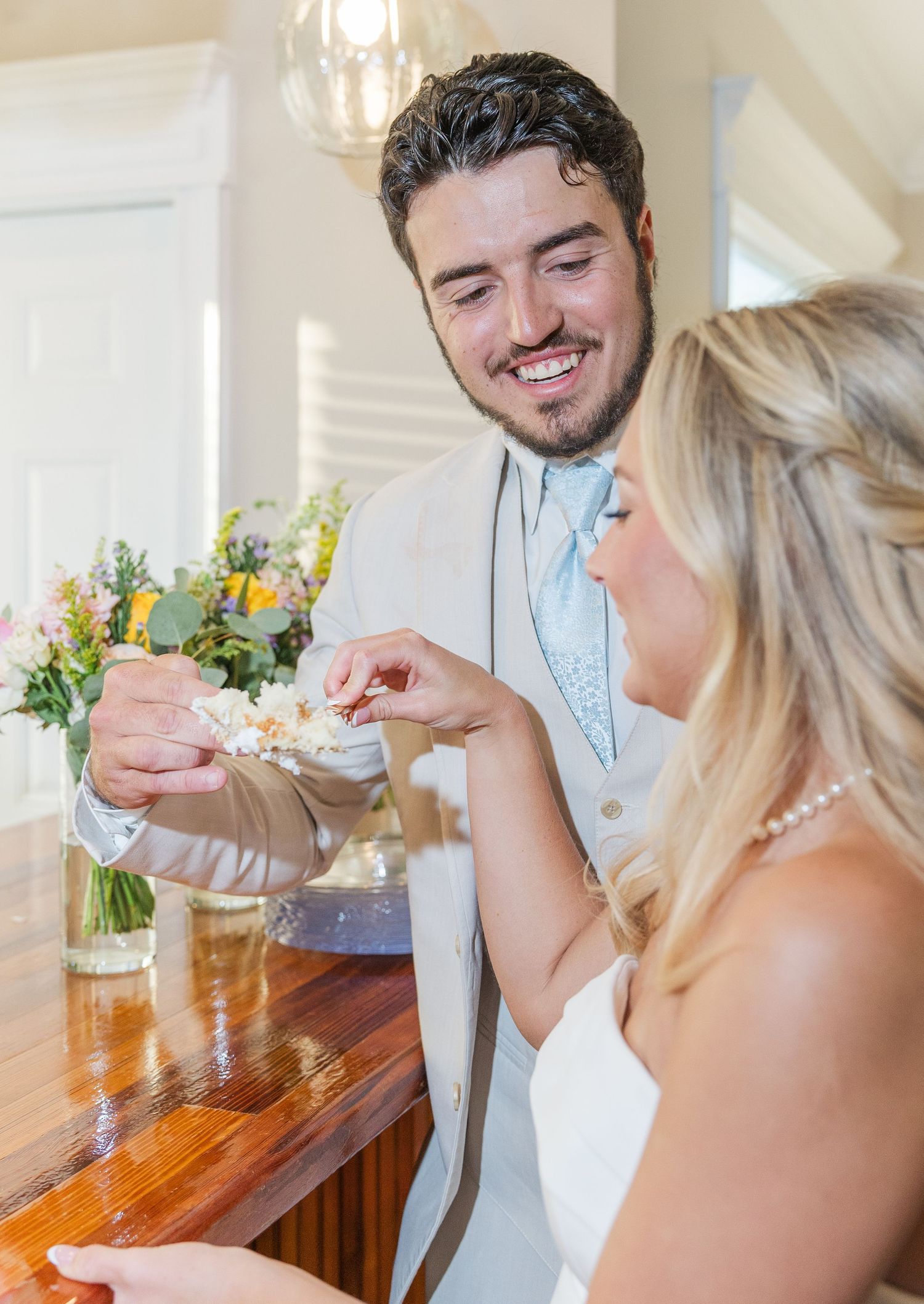 Bride and groom sharing wedding cake at reception, groom smiling in light gray suit with blue tie.