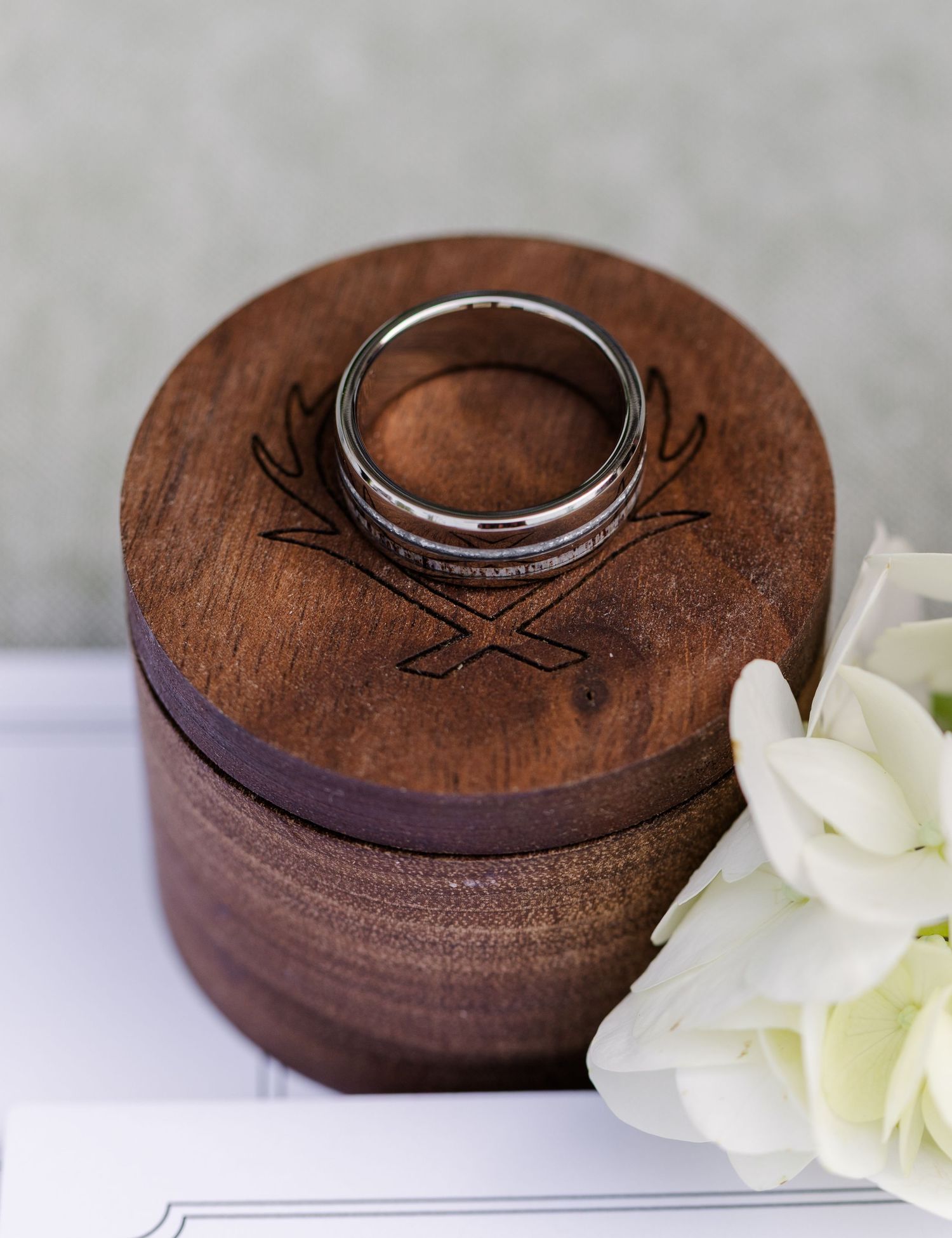 Silver wedding band resting on a round engraved wooden ring box with white flowers beside it.