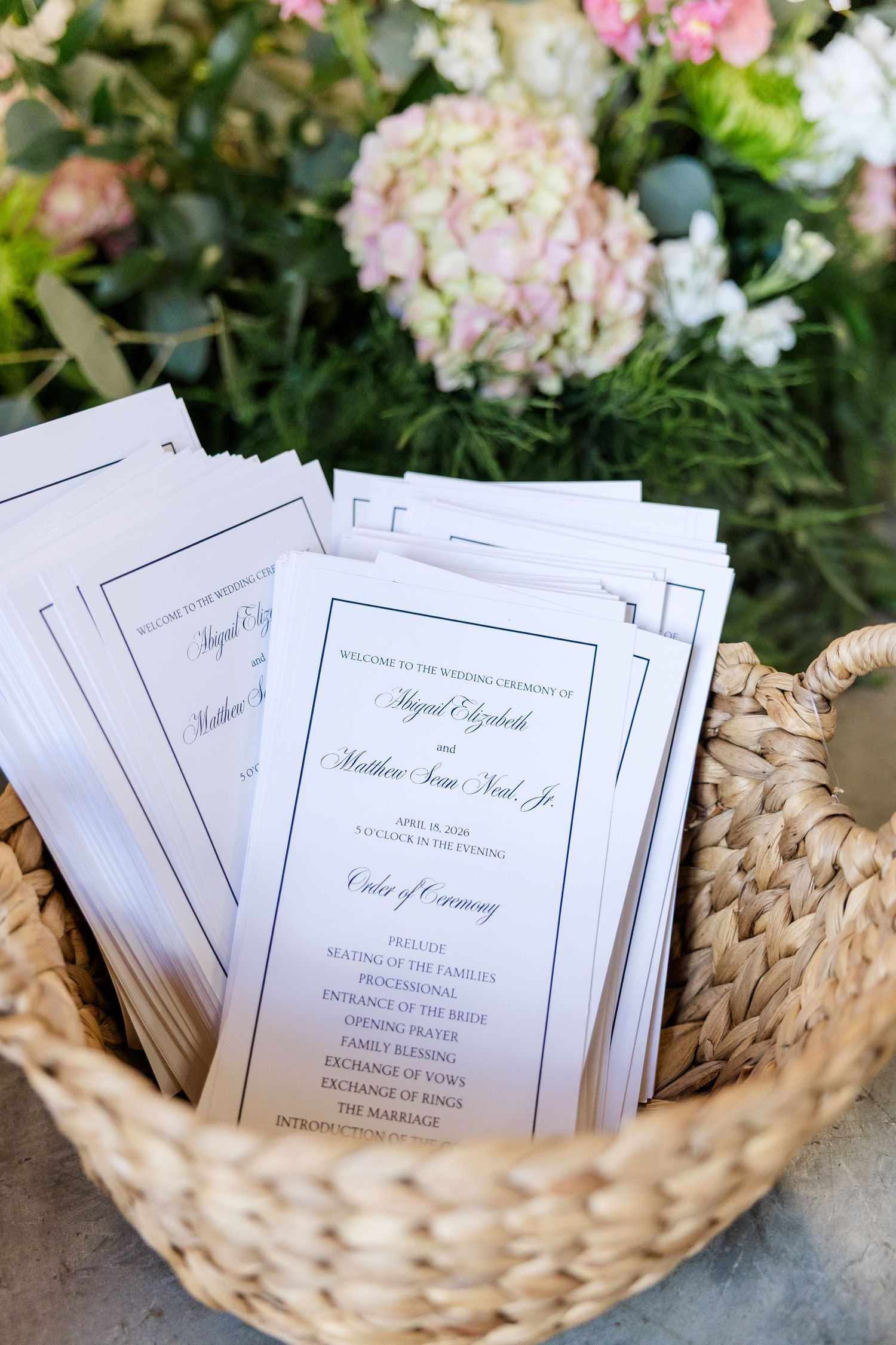 Wedding ceremony programs displayed in a wicker basket with pink hydrangeas in the background.