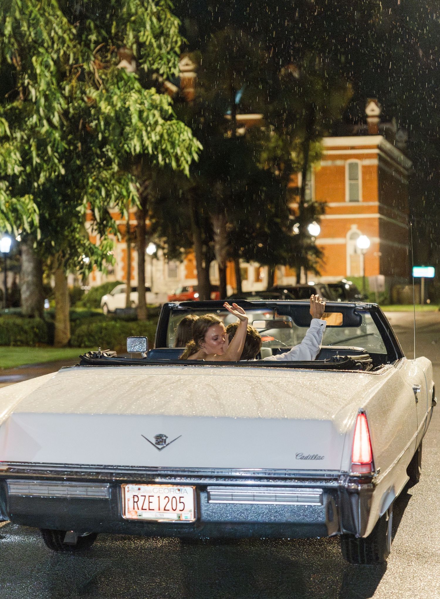 Newlyweds celebrate in vintage Cadillac convertible driving away in the rain at night.