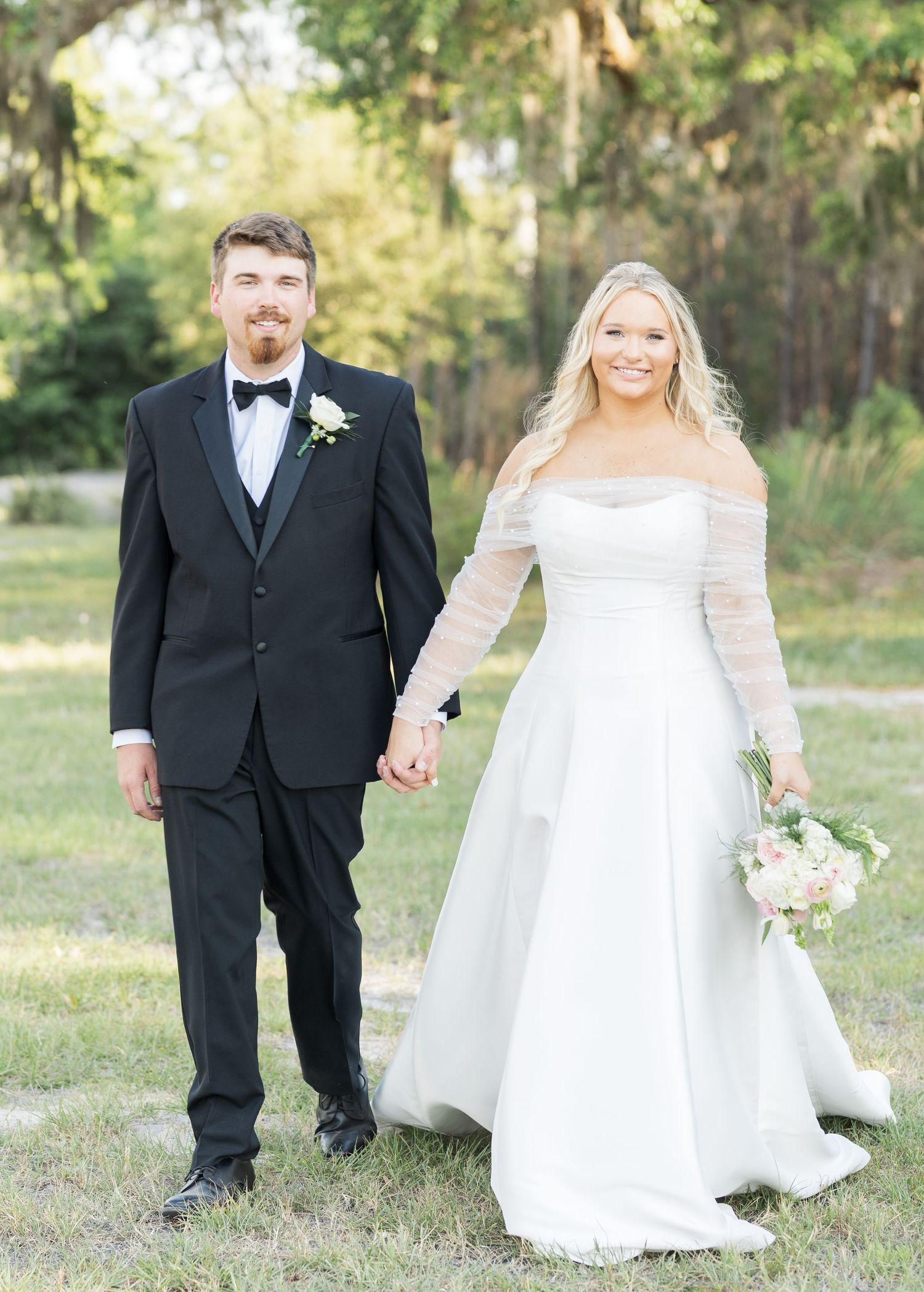 Bride and groom holding hands outdoors, she in an off-shoulder white gown, he in a black tuxedo with bow tie.