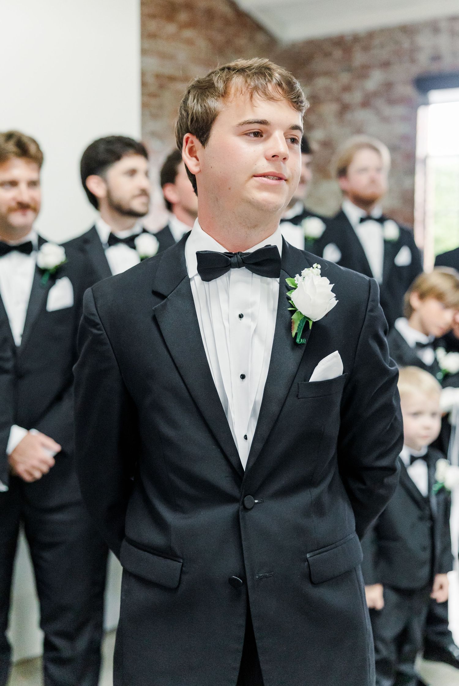 Groom in black tuxedo with white boutonniere stands at altar during wedding ceremony.