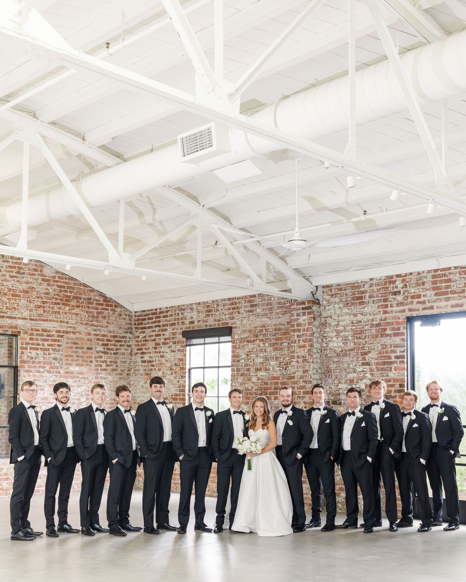 Bride and groom pose with large groomsmen party in black tuxedos inside rustic brick wedding venue.