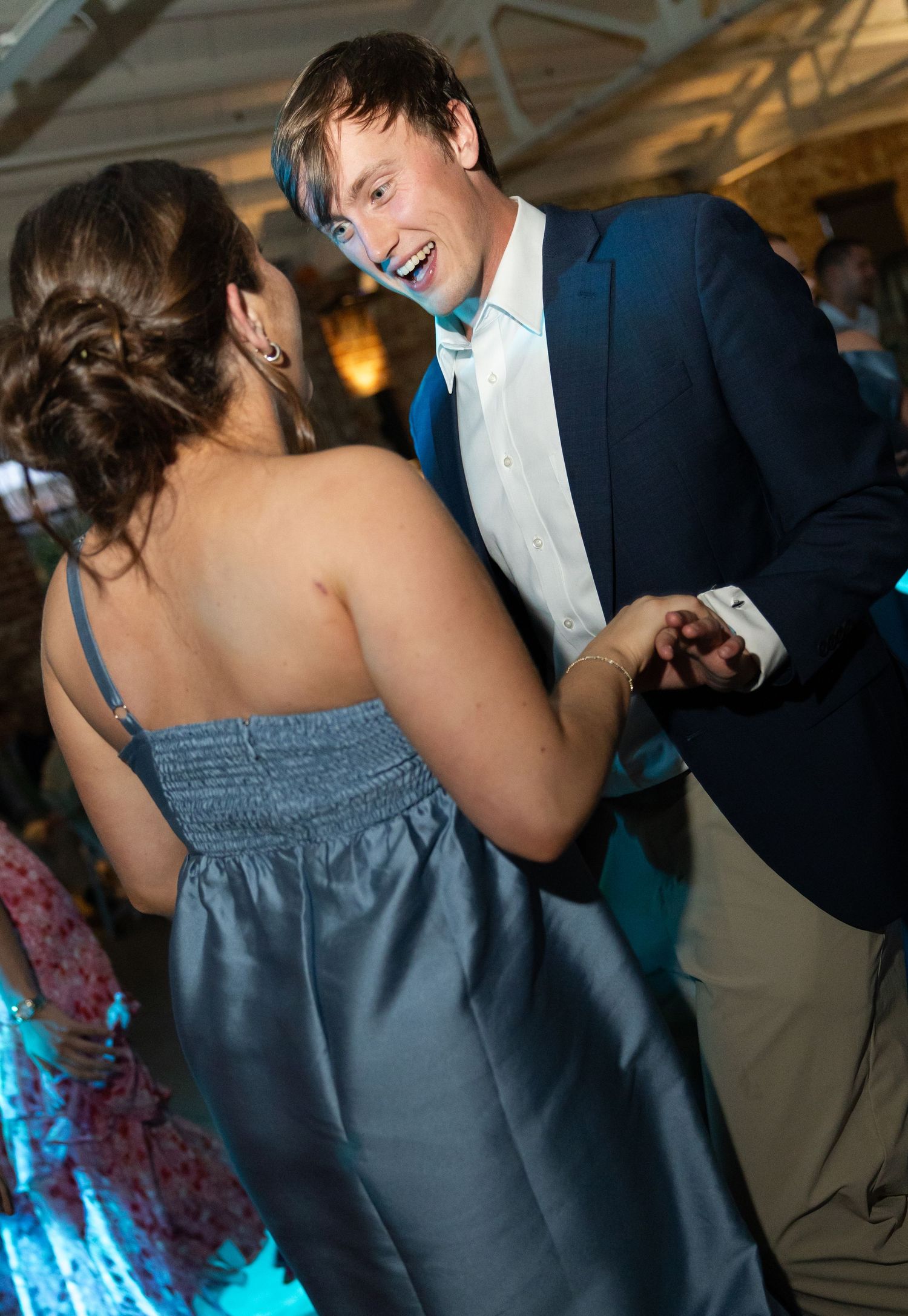 Smiling groomsman in navy suit dances with bridesmaid in blue dress at wedding reception.