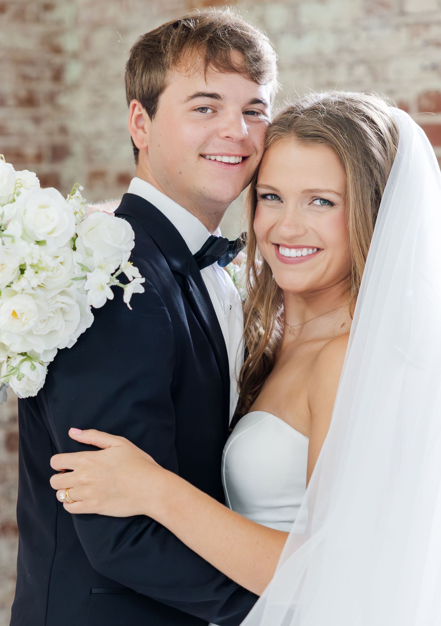 Happy bride and groom share a close romantic portrait pose, bride in strapless gown with veil, groom in black tuxedo.