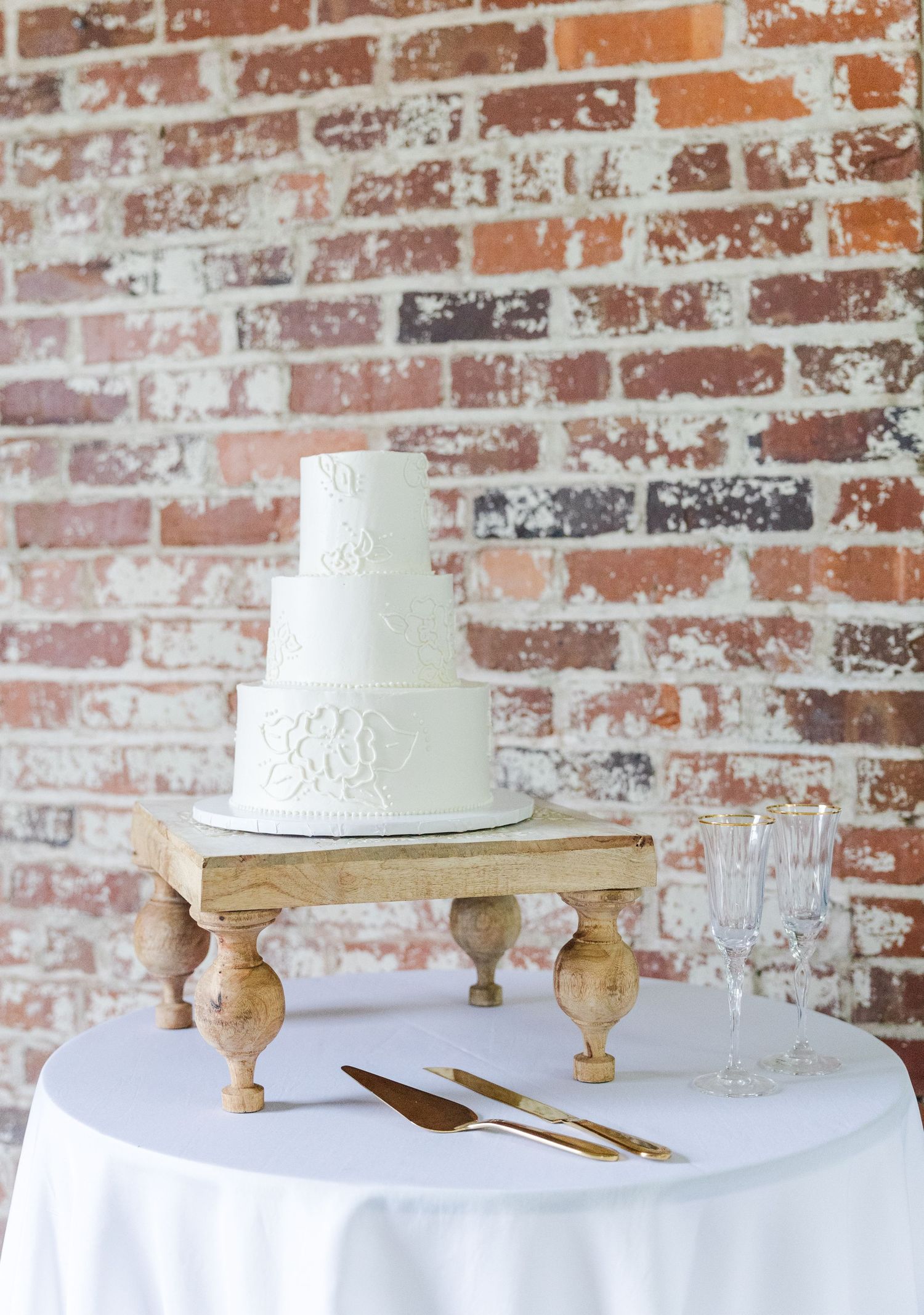 Three-tier white wedding cake with floral details on a wooden stand against exposed brick wall.