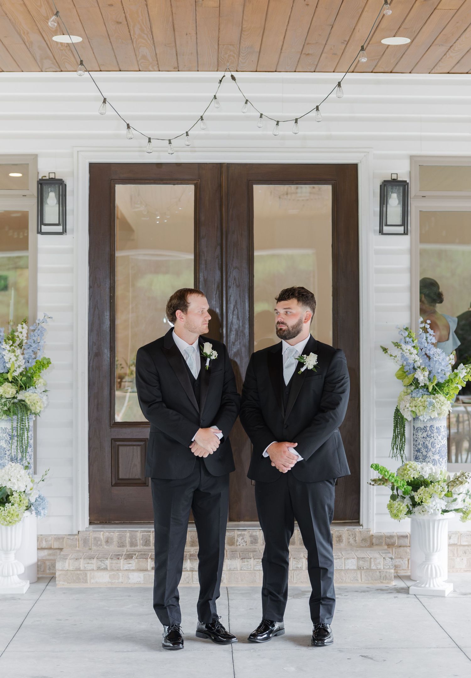 Two groomsmen in black suits with boutonnieres standing before wooden double doors at wedding venue.