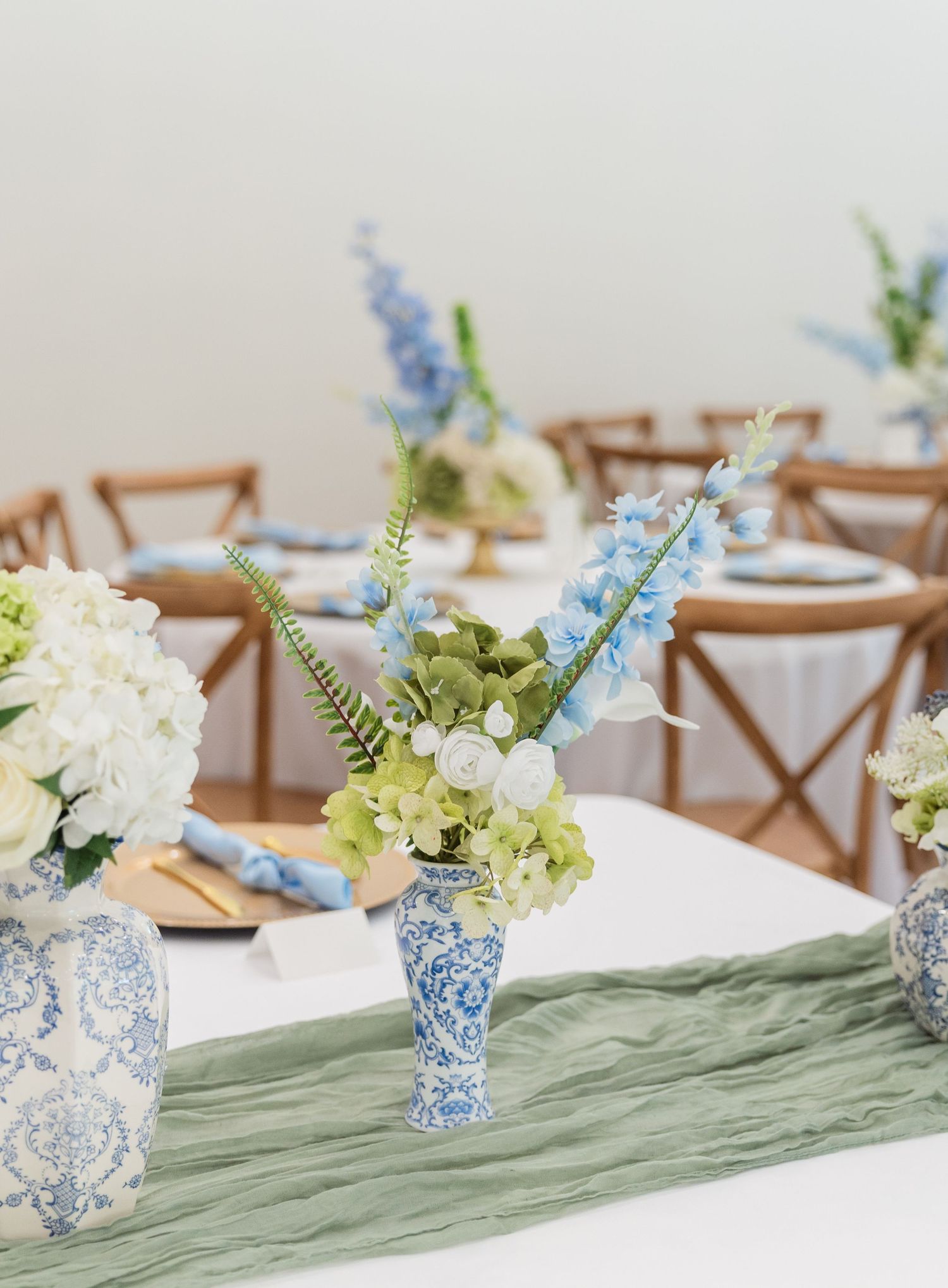 Blue and white chinoiserie vases with hydrangeas and delphinium on sage green table runner at wedding reception.