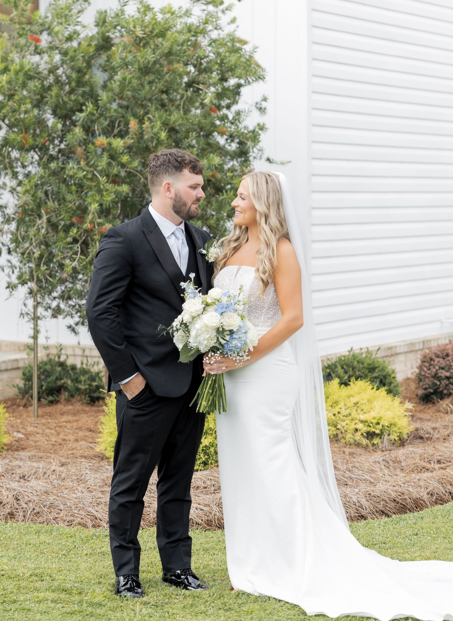 Bride and groom standing together outdoors holding a white and blue bouquet beside a white barn wall.