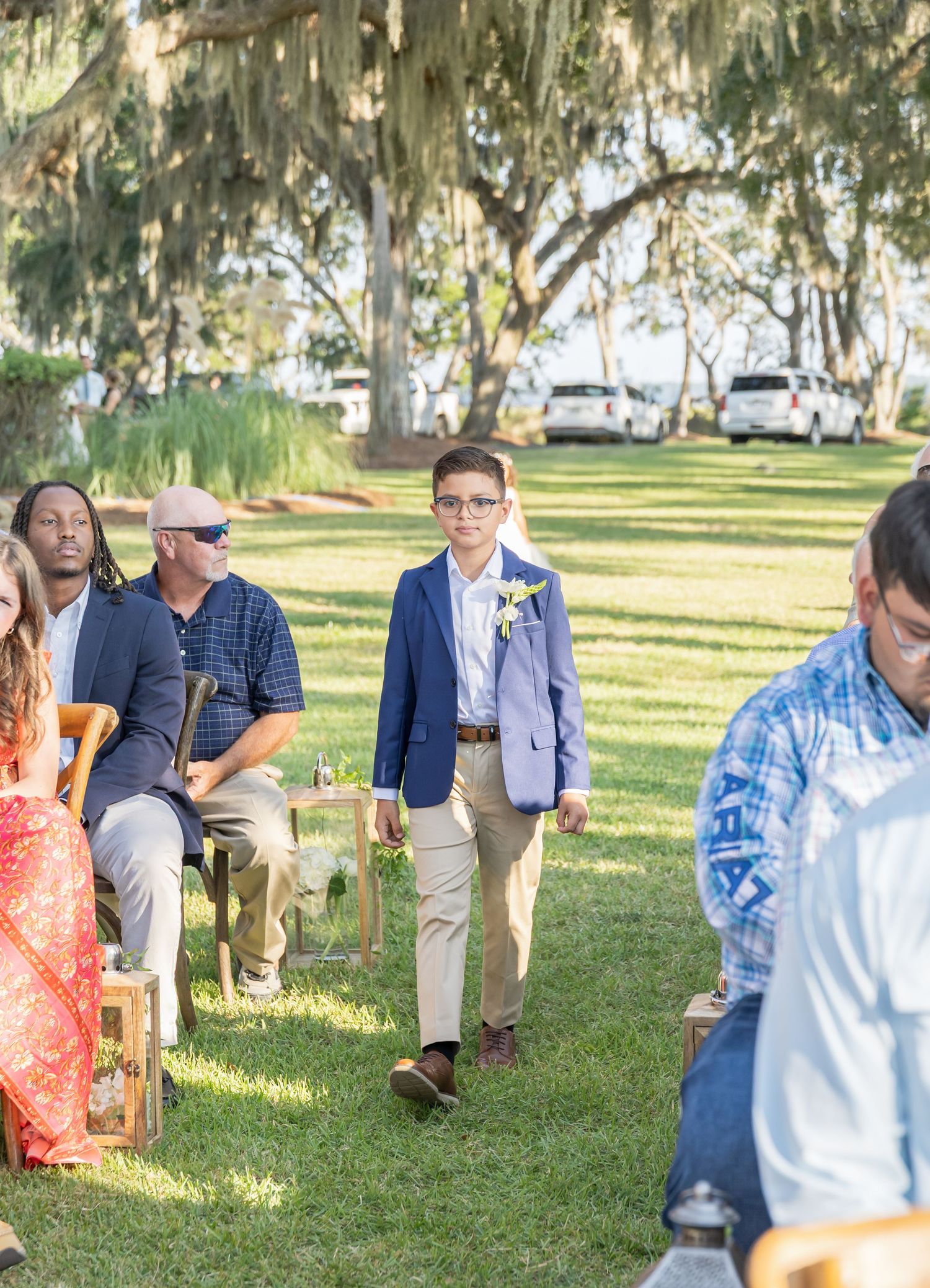Guests gather on a sunny lawn with Spanish moss-draped trees during an outdoor wedding ceremony.