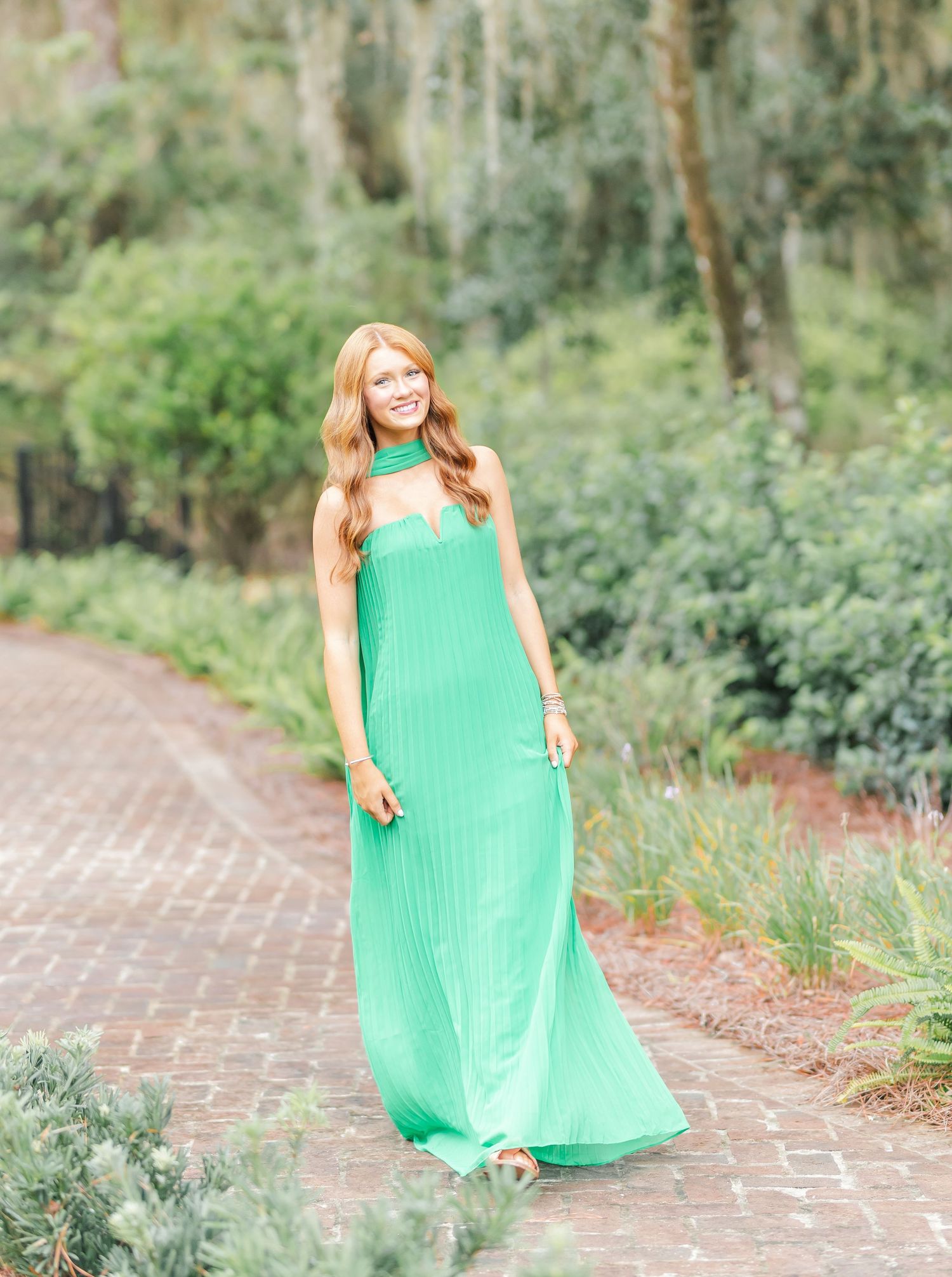 A woman in a flowing mint green maxi dress poses gracefully on a brick pathway surrounded by lush garden foliage.
