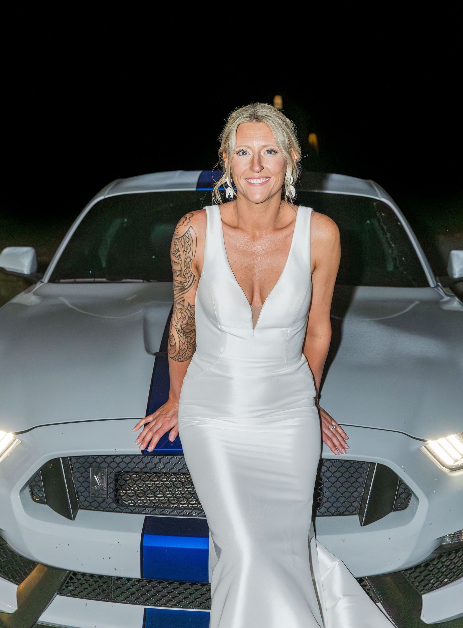 Bride in sleek white gown with sleeve tattoo poses on a silver Ford Mustang GT at night.