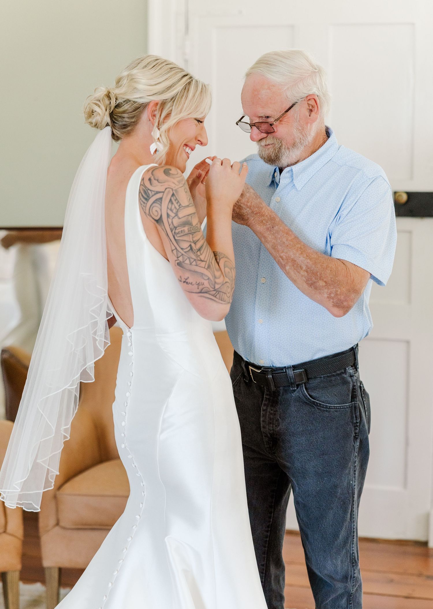 Older man helping smiling tattooed bride with her wedding dress and veil.