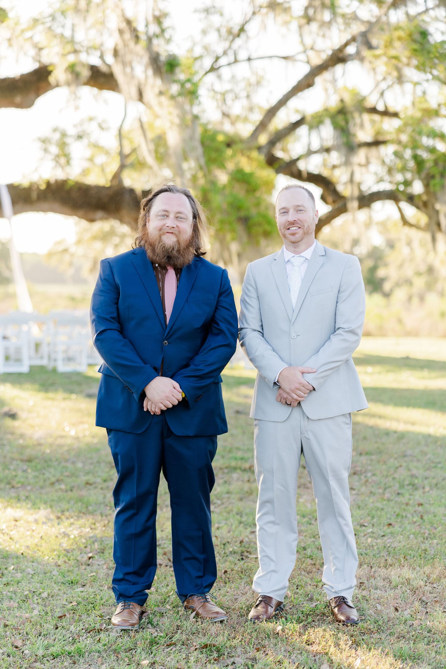 Two groomsmen in navy and gray suits smile outdoors near Spanish moss trees and ceremony chairs.