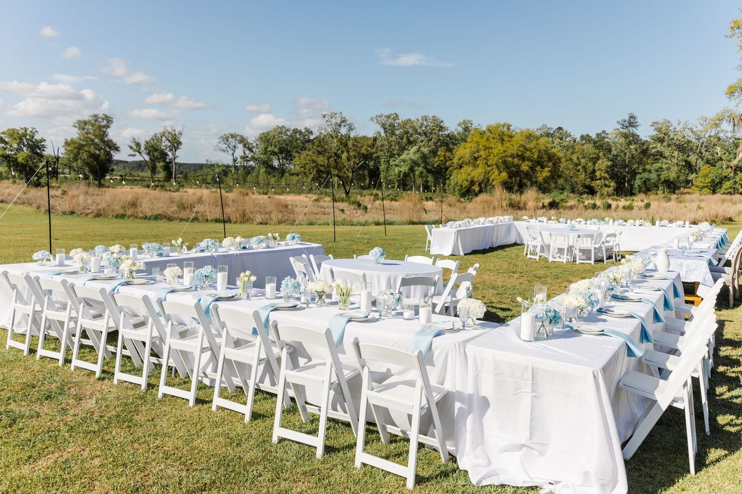 Outdoor wedding reception setup with long white-draped tables and white folding chairs on a green lawn.