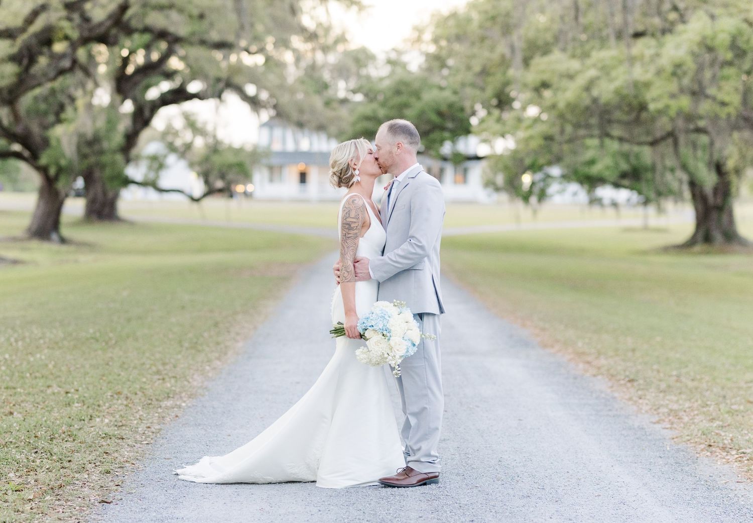 Bride and groom sharing romantic kiss on tree-lined driveway at golden hour during wedding day.