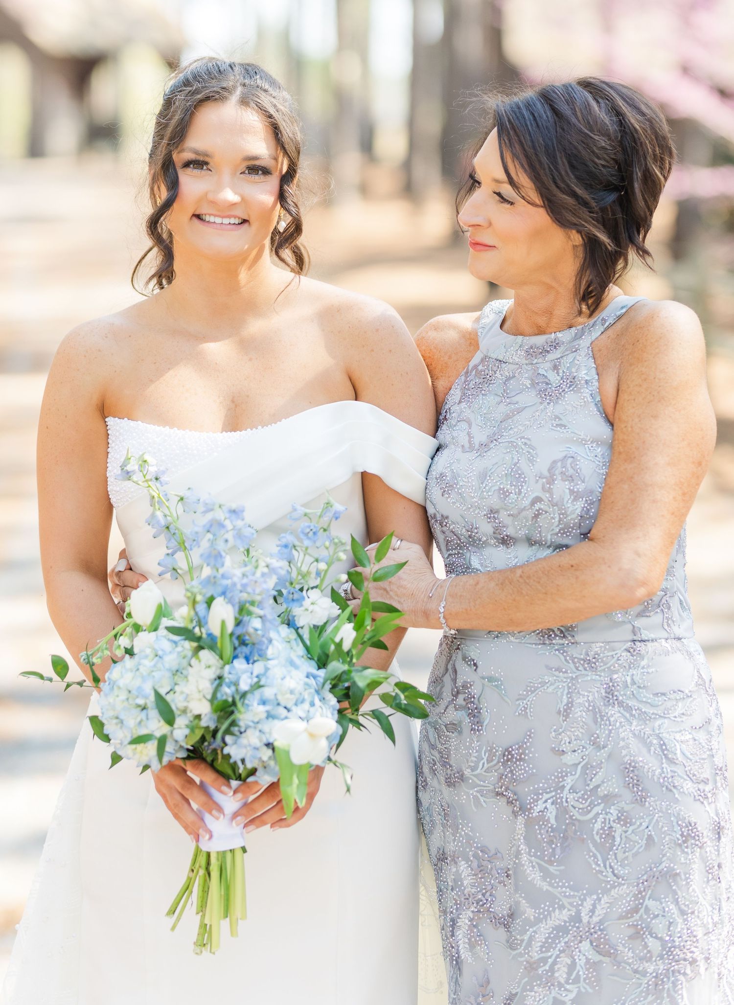Bride in strapless gown holding blue hydrangea bouquet posing with mother in lavender lace dress.