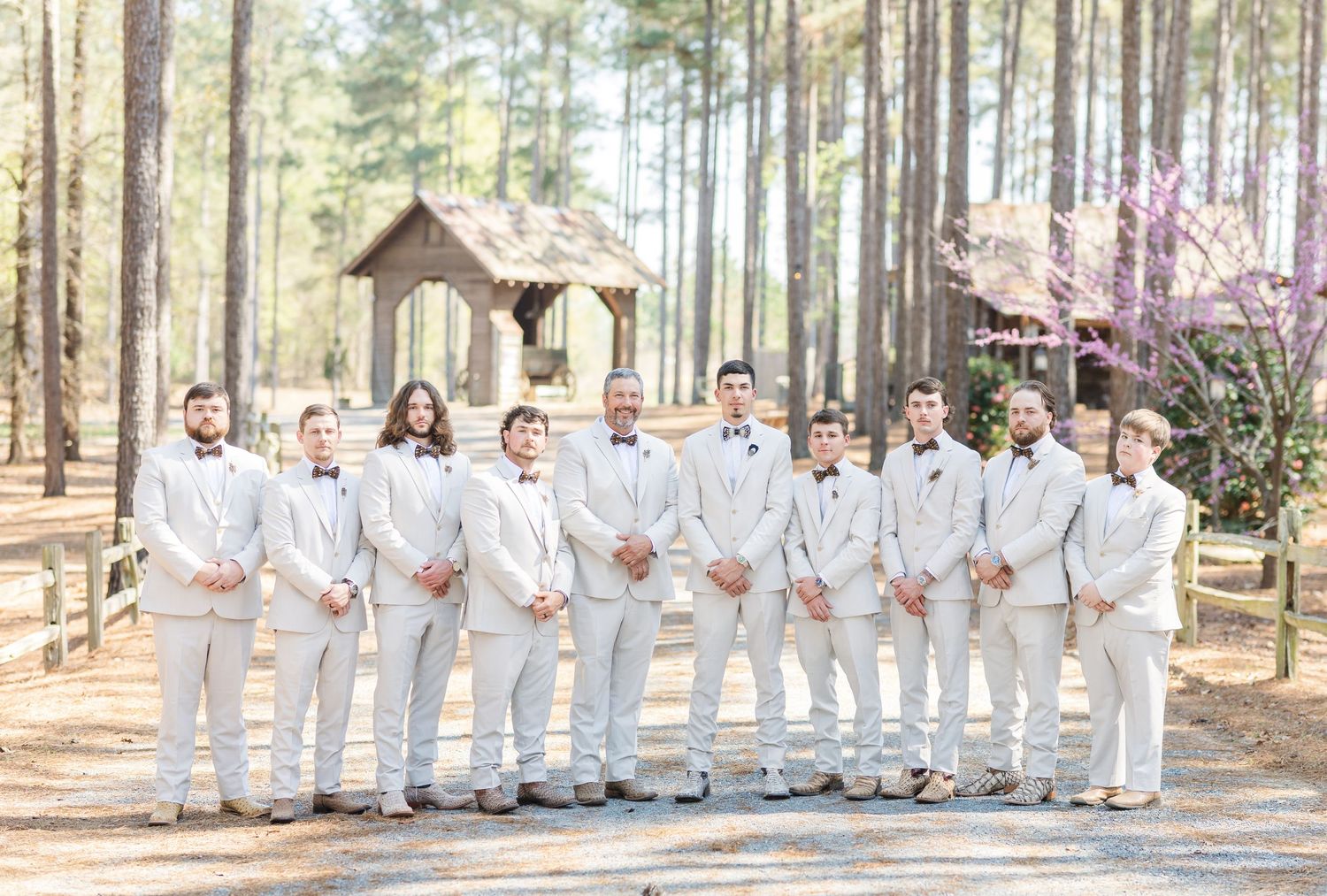 Groomsmen in white suits pose outdoors at a woodland wedding venue with pine trees and a rustic gazebo.